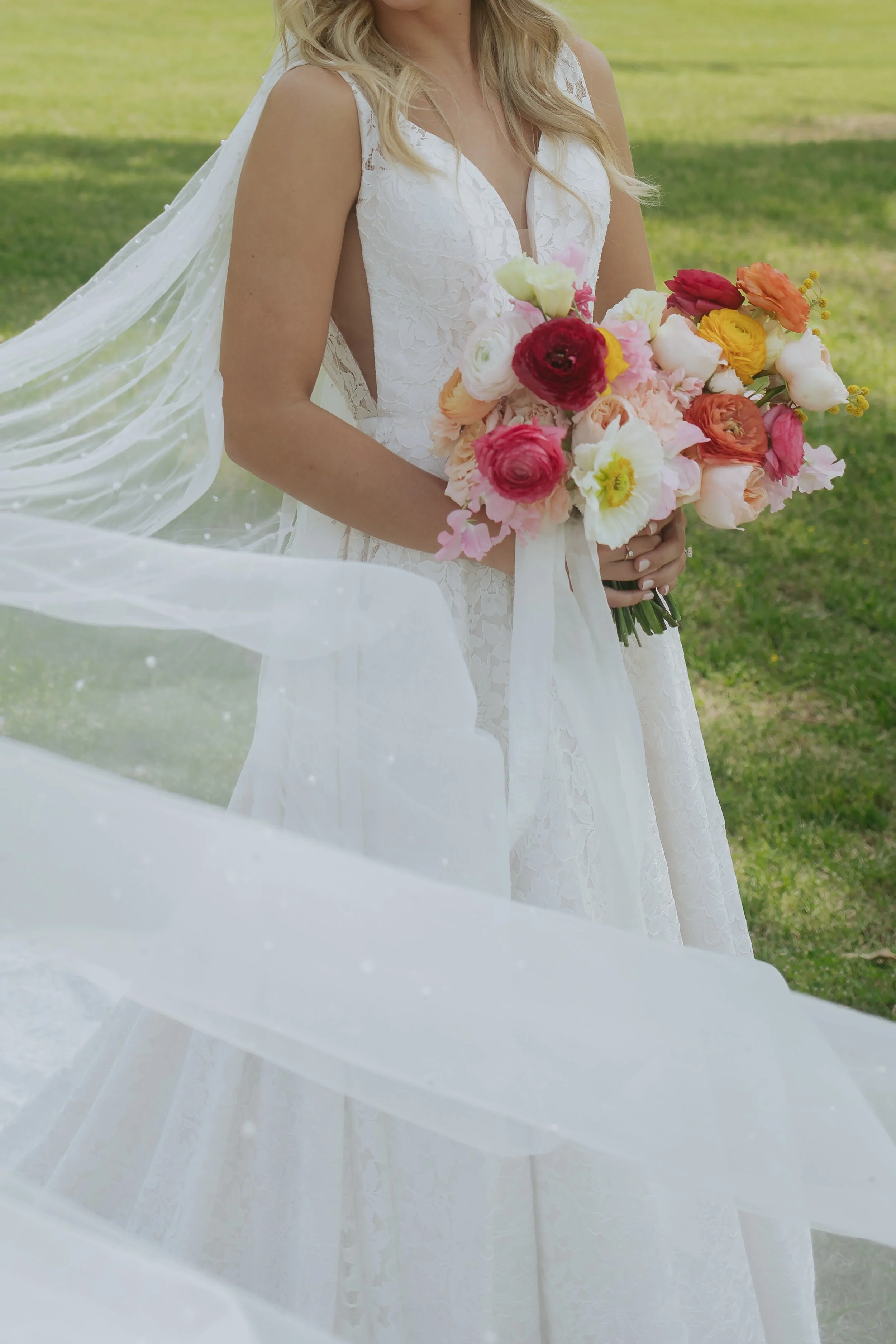 A bride in a white lace wedding dress holding a colorful bouquet of flowers, outdoors on grass.
