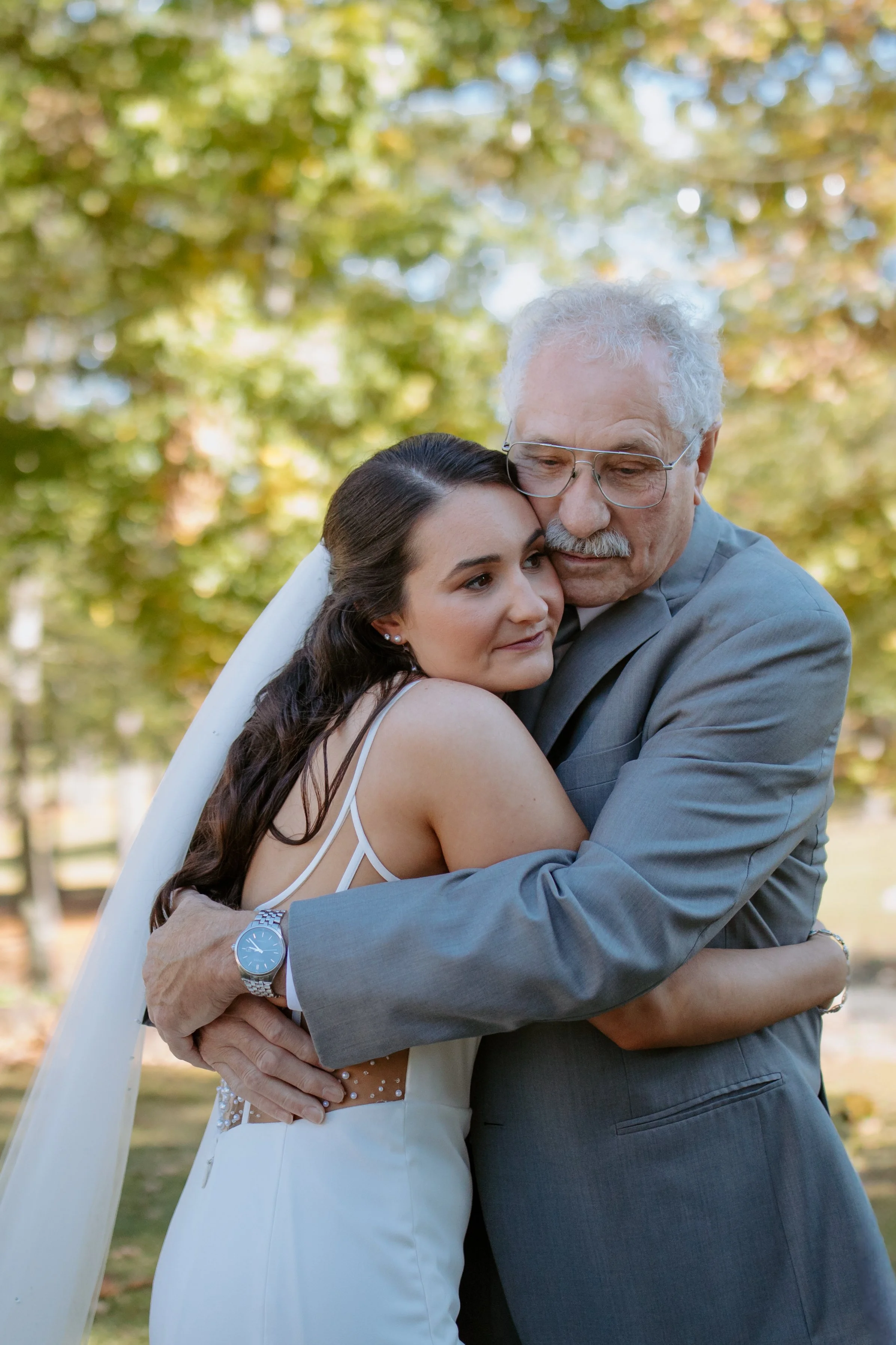 A bride hugging an older man, likely her father, outdoors during daytime with autumn trees in the background.