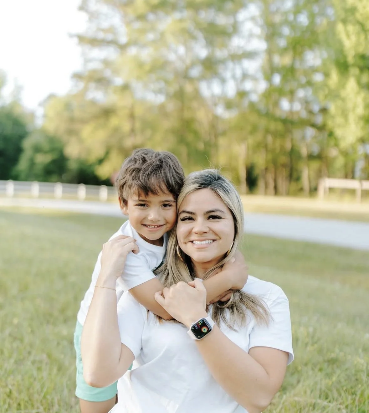 A woman with a smartwatch on her wrist holding a young boy, both smiling outdoors in a grassy area with trees in the background.