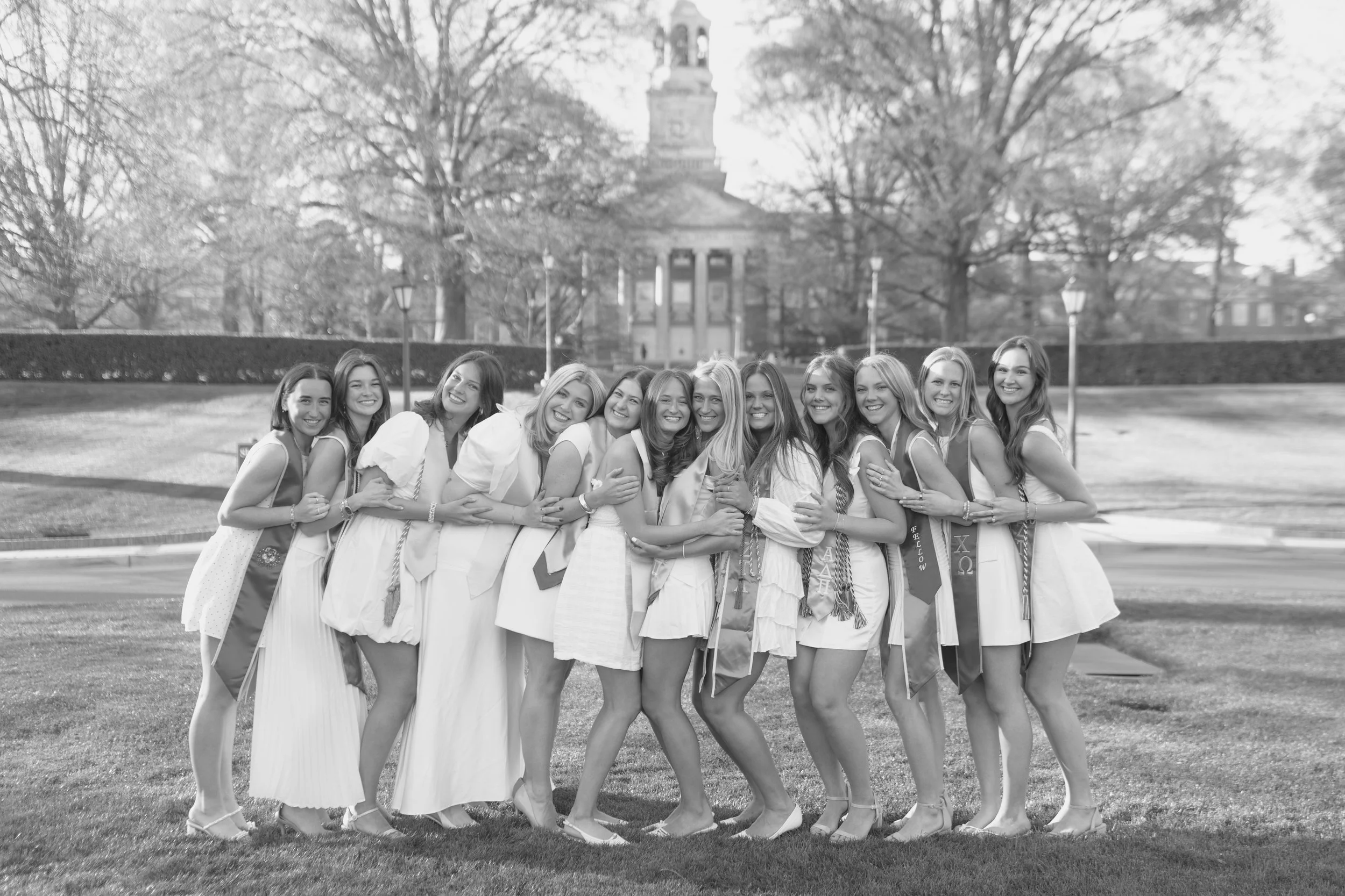 A group of young women in white dresses and sashes, celebrating outdoors on a lawn with trees and a building in the background.