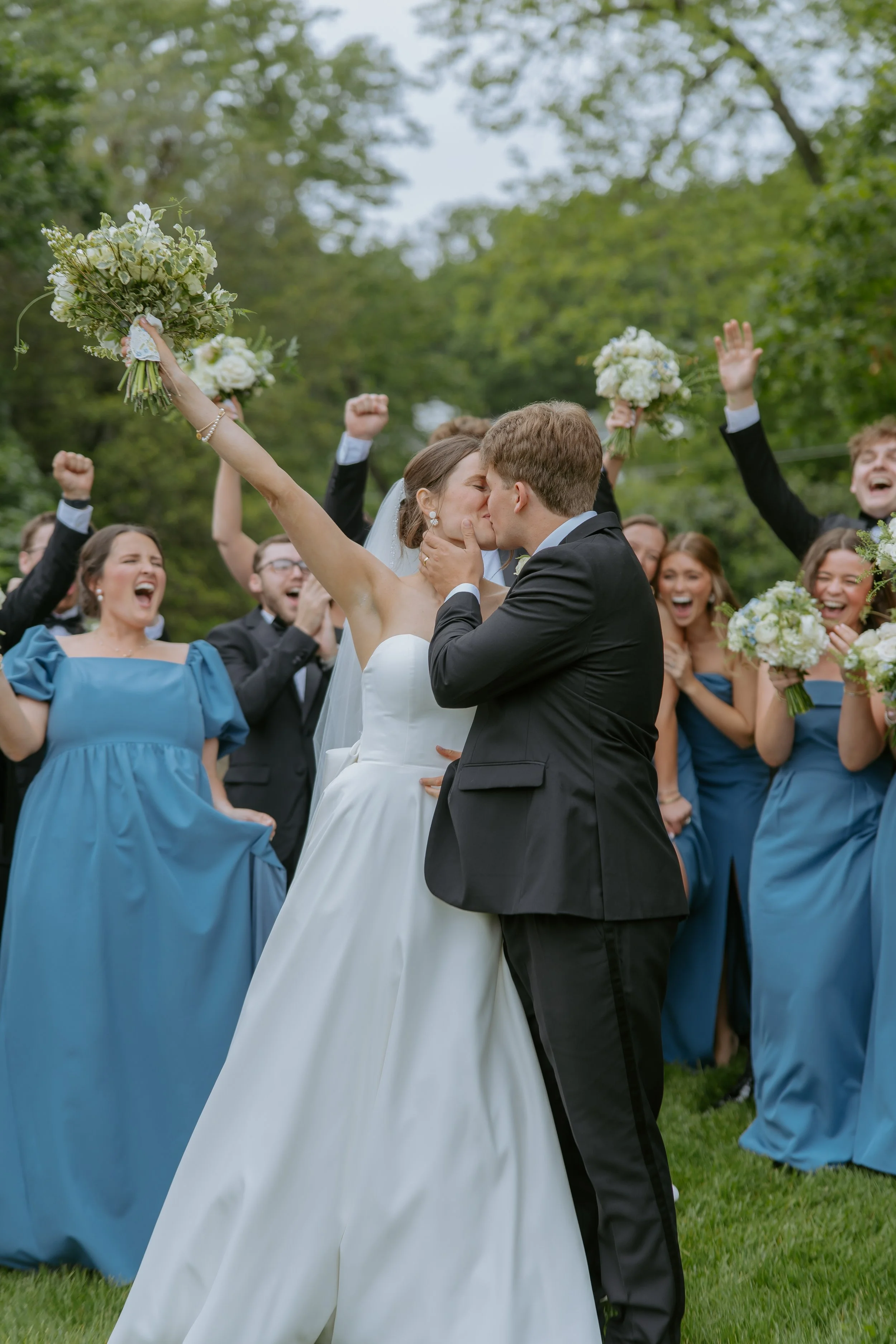 A bride and groom kiss as friends celebrate around them, with the bride holding a bouquet, outside in a green, wooded area.