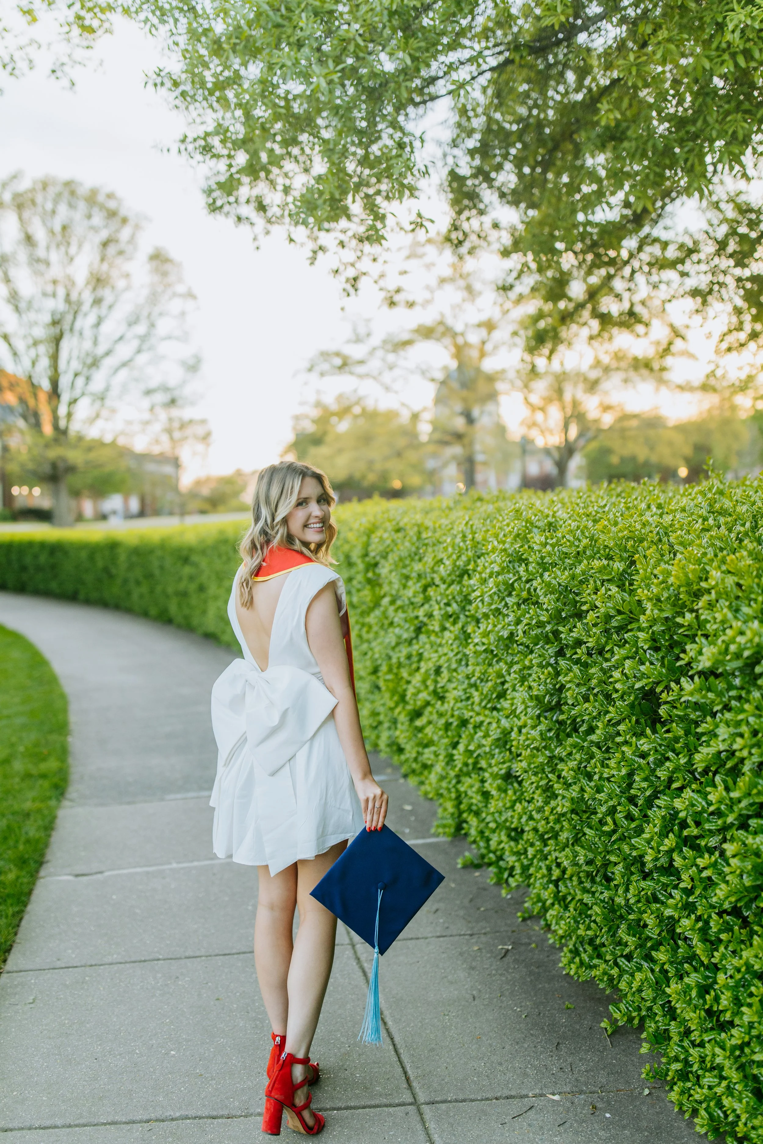 A young woman in a white graduation dress with red accents and red high heels, holding a navy blue graduation cap, stands on a sidewalk next to a green hedge, smiling at the camera, with trees and a setting sun in the background.