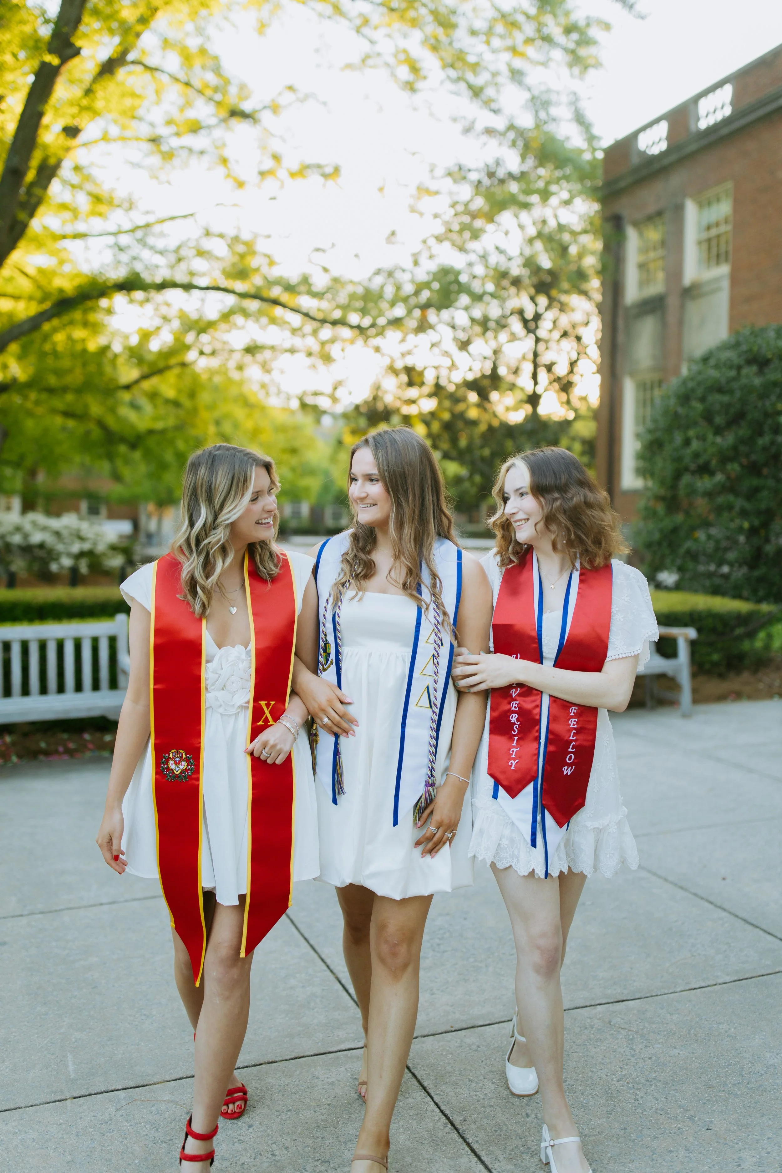 Three young women in white dresses and graduation sashes walking outdoors in a park with trees and a brick building in the background, smiling and talking.