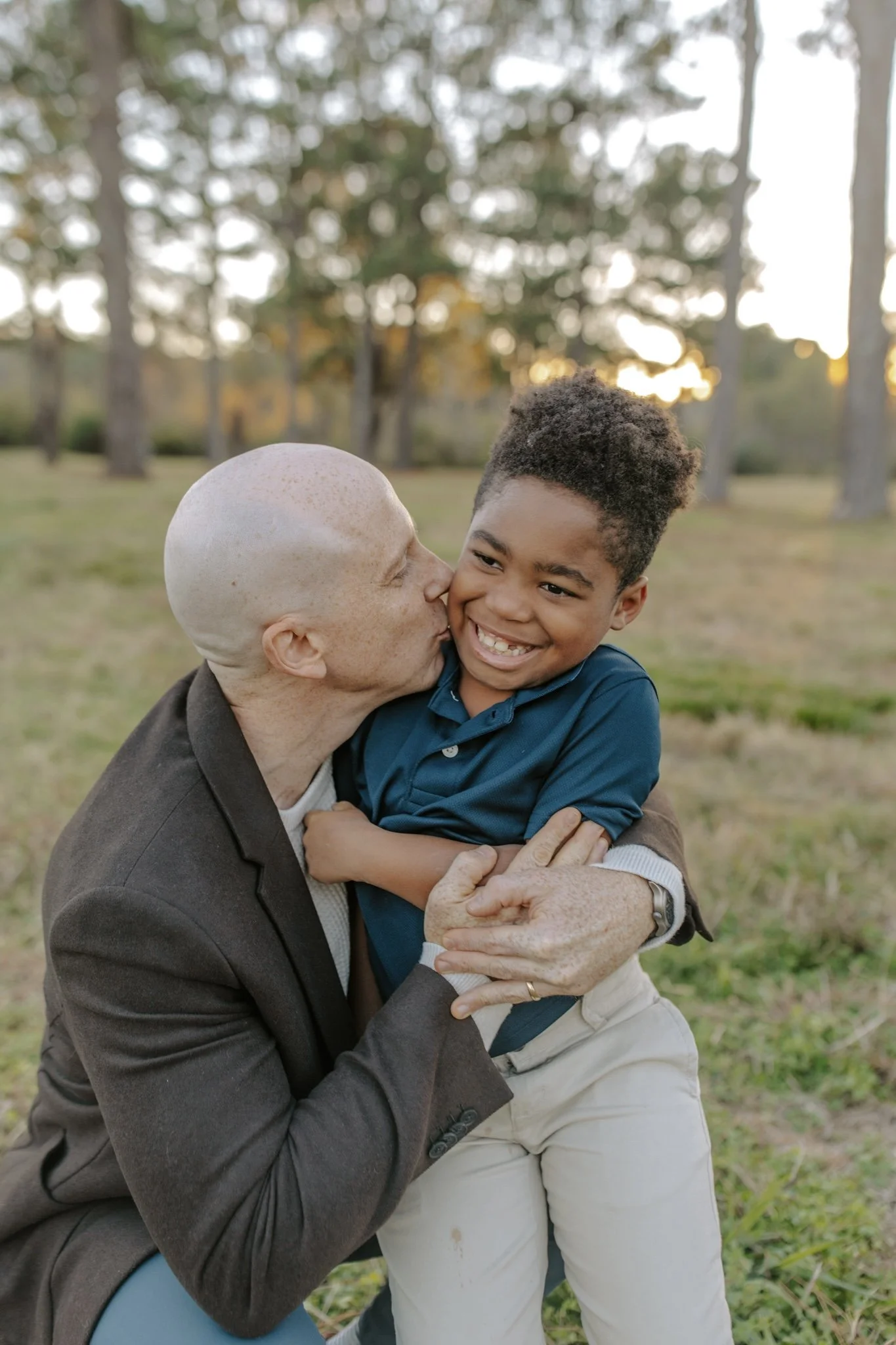 An elderly man with a bald head and light skin kissing a young boy with dark curly hair and dark skin on the cheek, both smiling happily outdoors with trees and sunset in the background.