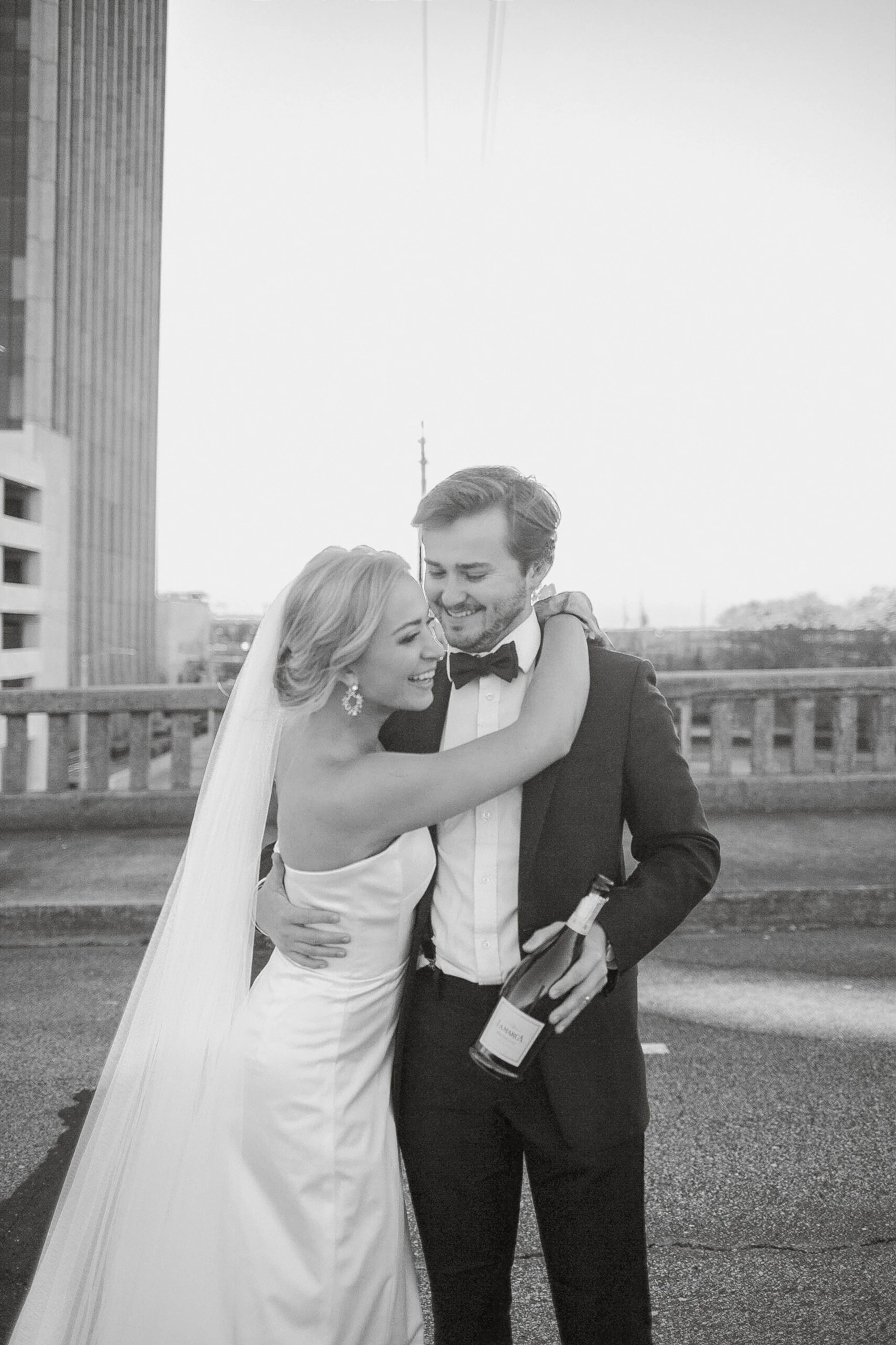 A bride and groom sharing an embrace on a rooftop, with the bride smiling and hugging the groom, who is holding a bottle of champagne, in a black and white photo.