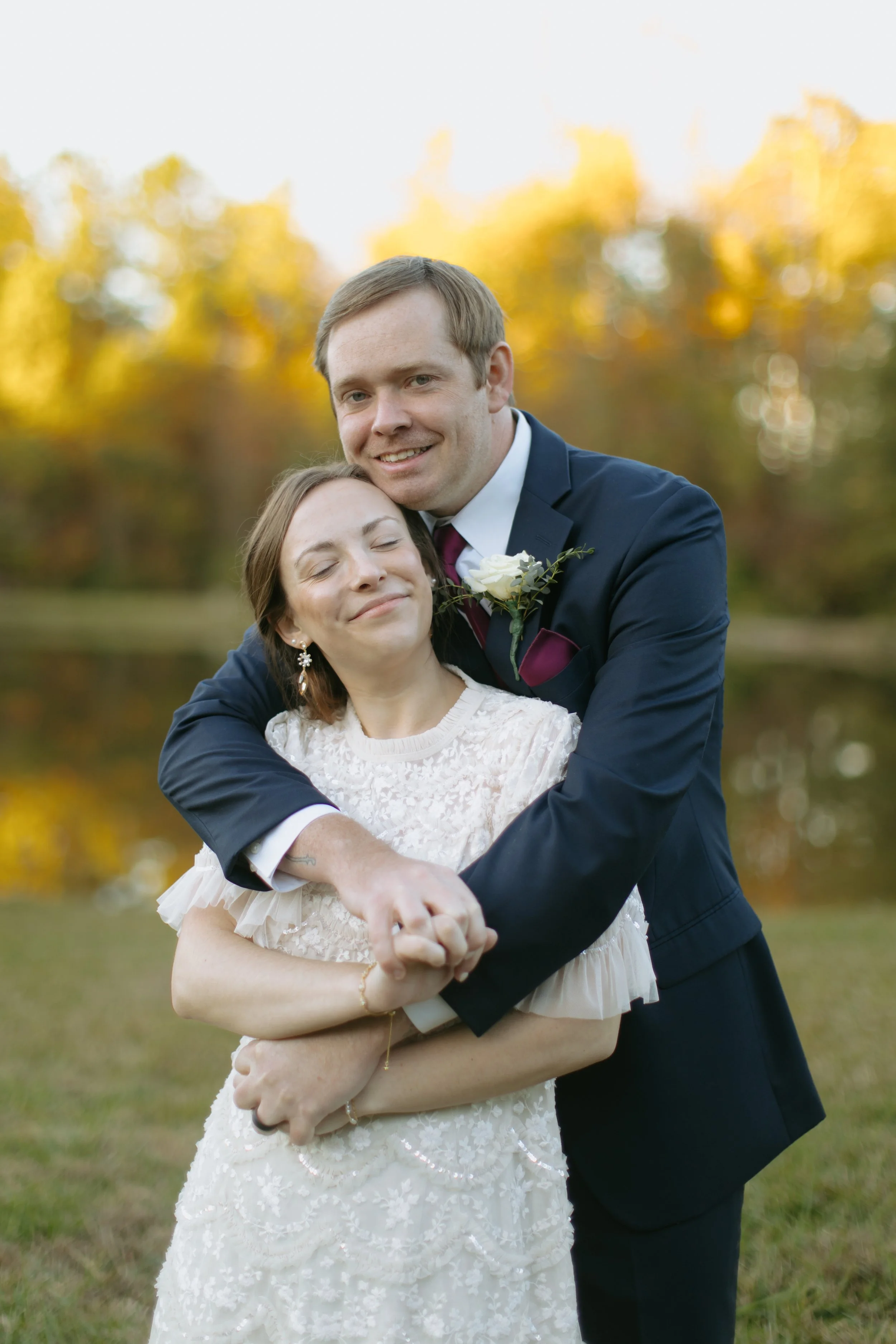A happy couple hugging outdoors during sunset, with warm autumn trees in the background. The man is dressed in a dark suit with a white shirt and a boutonniere, and the woman is wearing a white dress with lace details and earrings.
