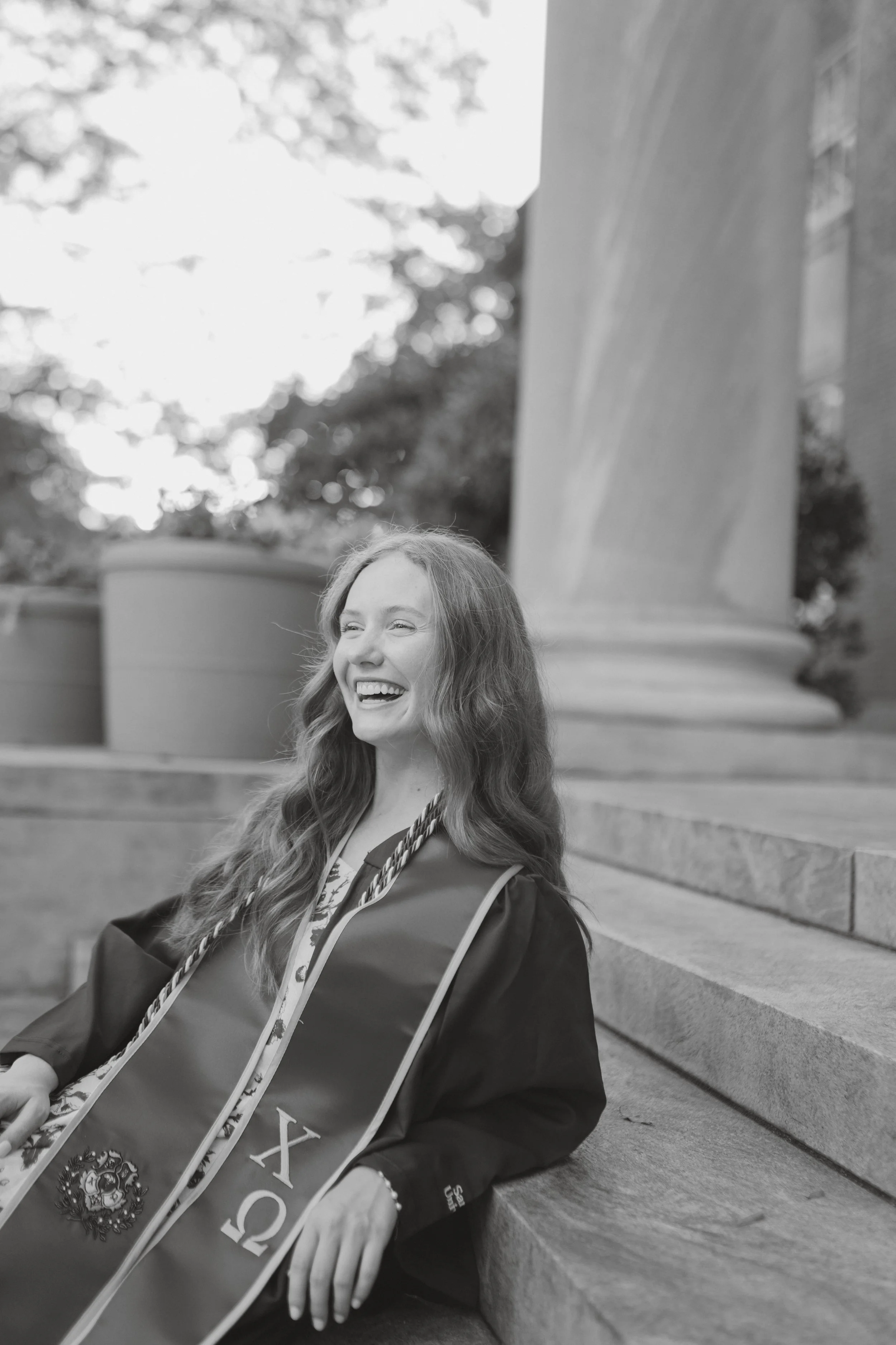 Black and white photo of a young woman in graduation gown and sash, sitting on steps outdoors, smiling and laughing.