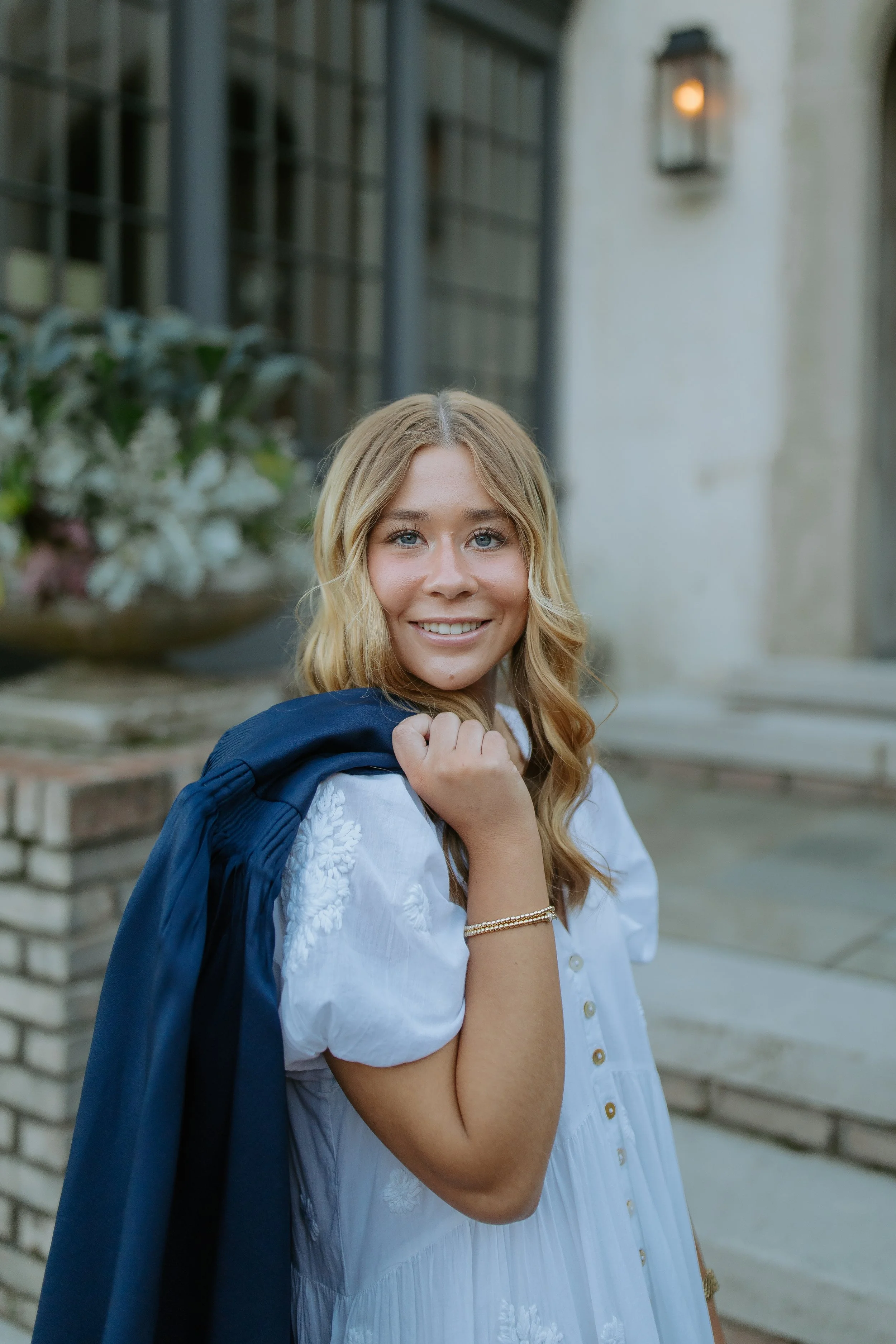 A young woman with blonde wavy hair smiling and holding a navy blue jacket over her shoulder, standing outside near a brick wall and a door with a lantern.