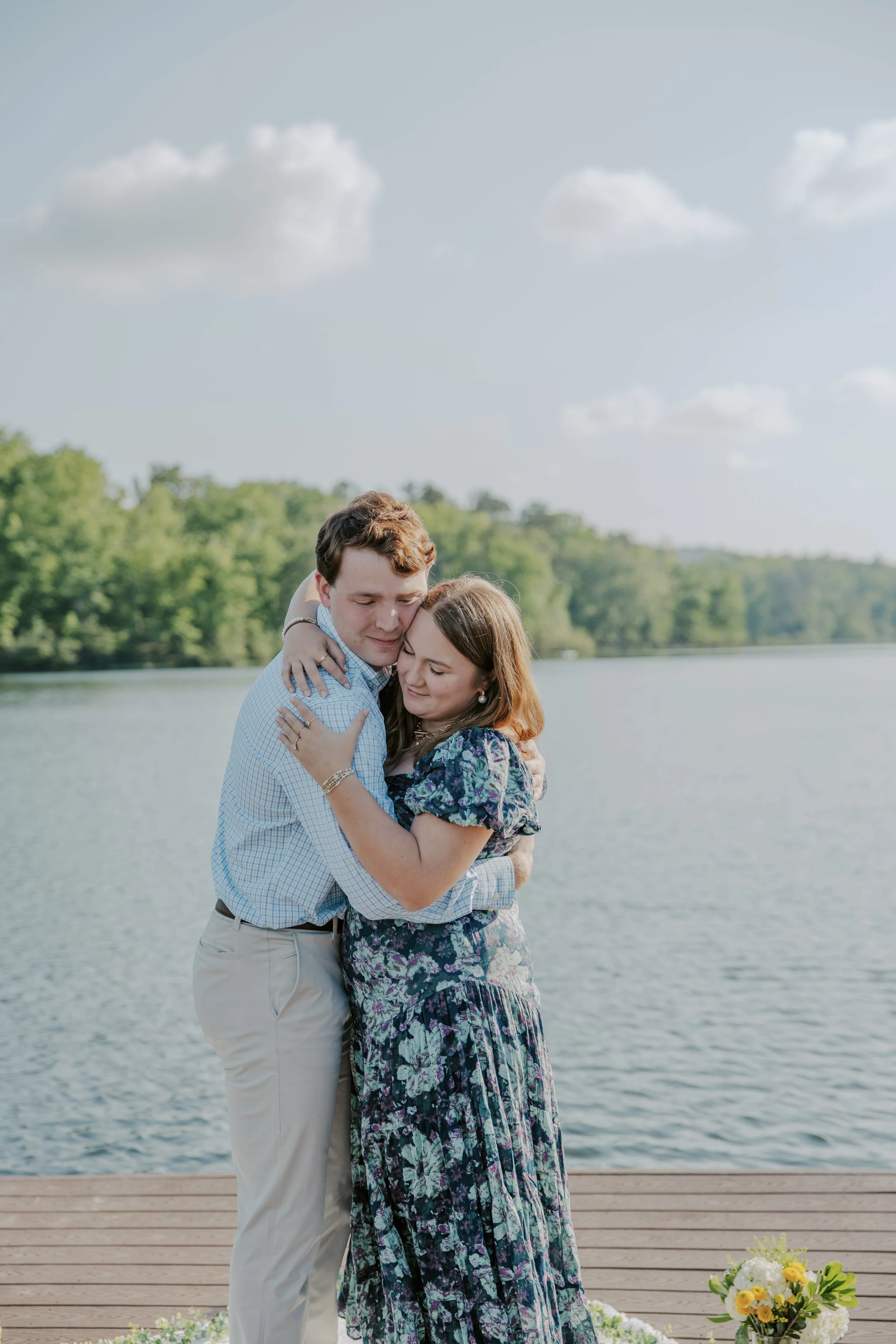 A couple hugging by a lake with trees in the background on a sunny day, with a floral arrangement on the dock.