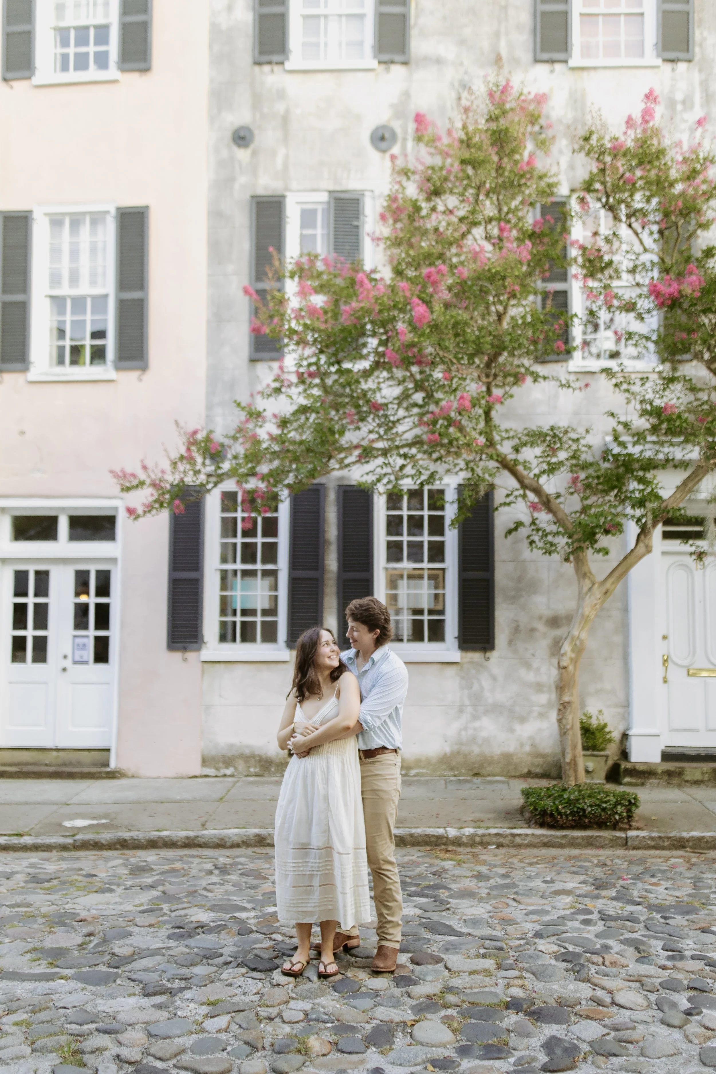 A young couple smiling and hugging on a cobblestone street with a beige building, pink blossoming trees, and black window shutters in the background.