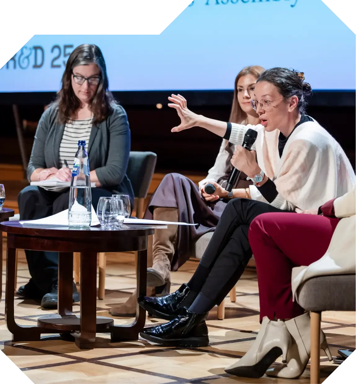 A panel discussion featuring three women seated, with one actively speaking into a microphone while gesturing.