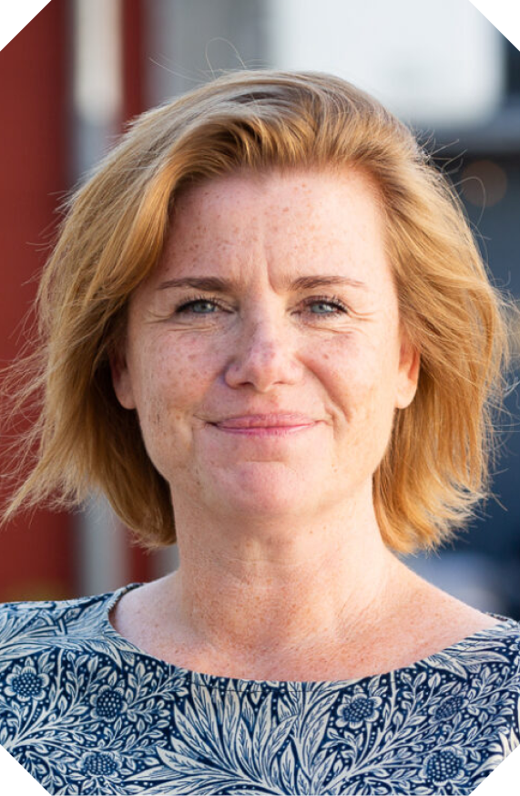 A woman with short red hair and blue eyes, smiling slightly, wearing a blue and white patterned top, outdoors with a blurred background.