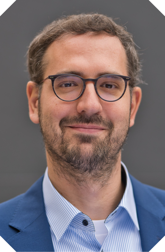 A man with glasses, a beard, and short curly hair is smiling and wearing a blue blazer and light blue shirt.