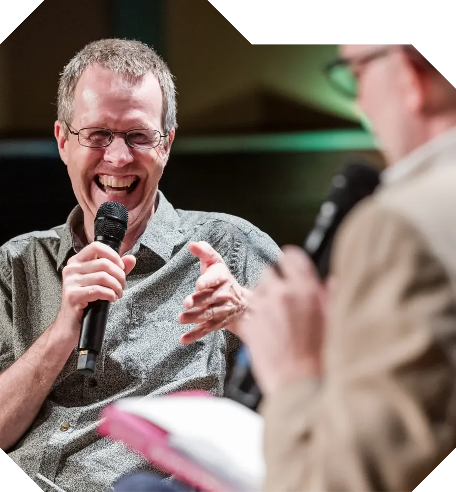 A man with glasses laughs while speaking into a microphone during a discussion, gesturing enthusiastically.