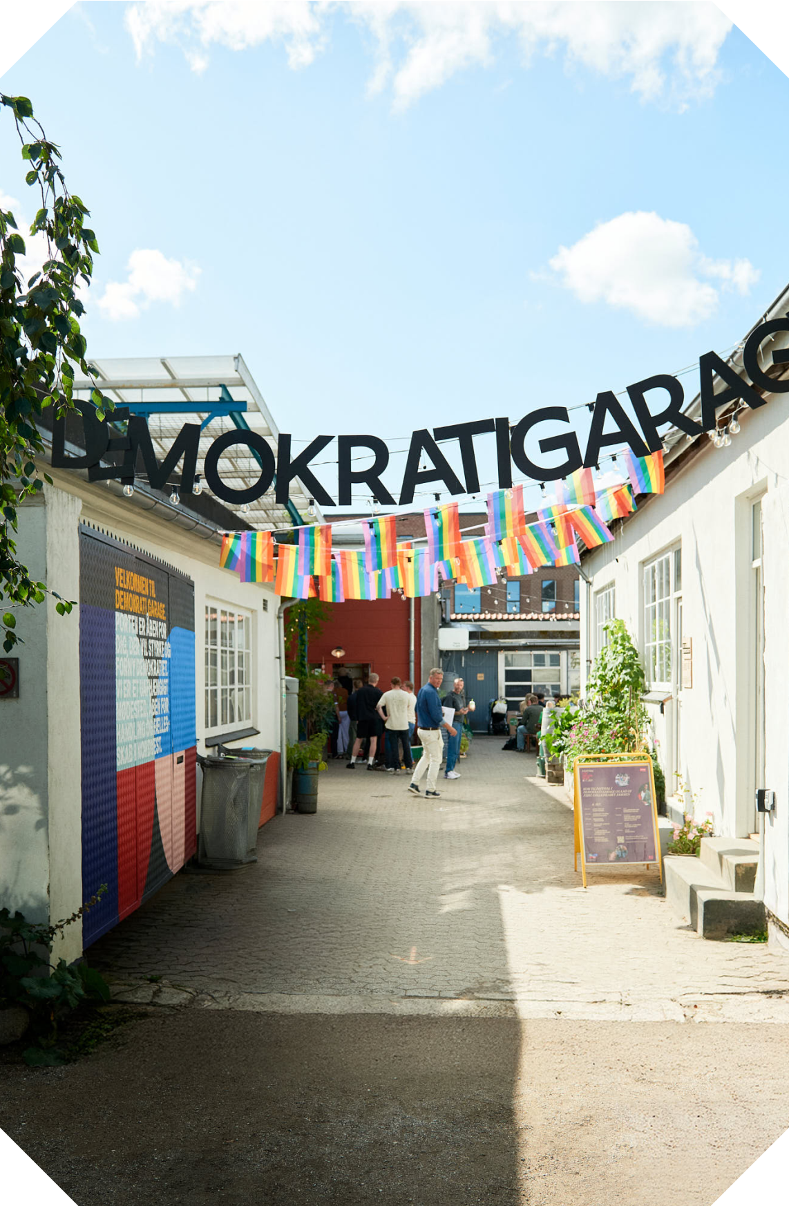 A street scene during a pride event with people gathered under a banner that reads 'DEMOKRATIGAR' and decorated with rainbow pride flags, a colorful poster on the left, and a small outdoor sign on the right, with bright blue sky and white clouds overhead.