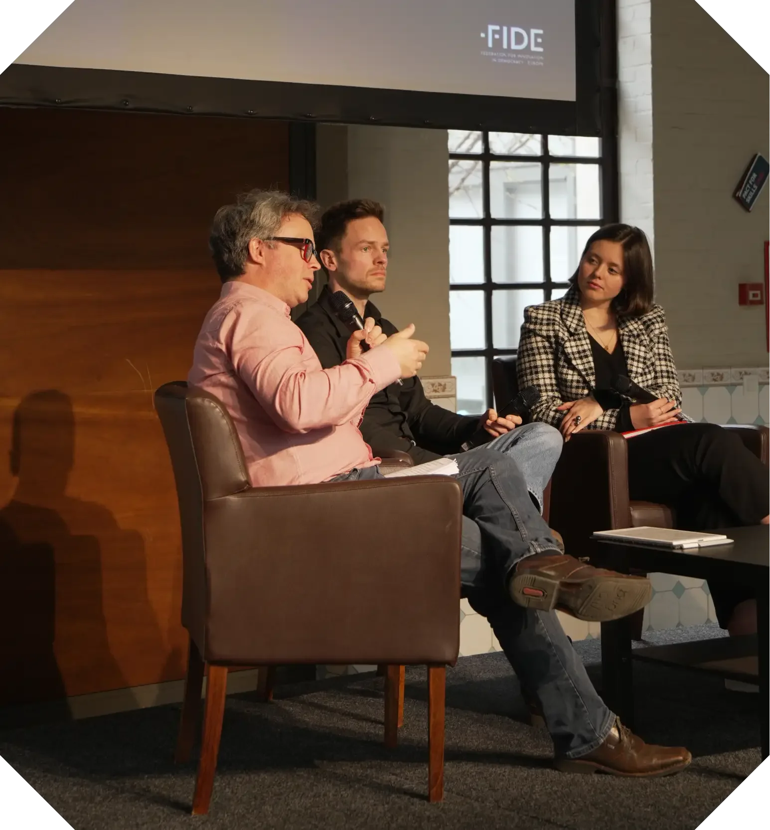 Three panelists engage in discussion at an event, with one speaking into a microphone while the others listen attentively.