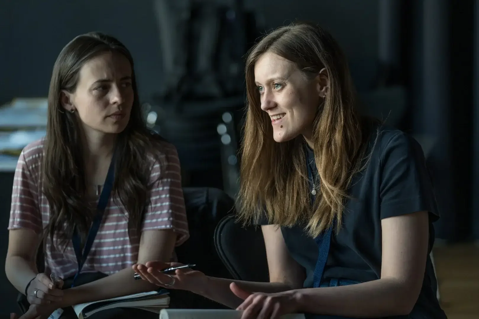 Two women sitting and talking in a dimly lit room, one with long dark hair and the other with long auburn hair. They have notebooks and pens, engaged in a conversation.