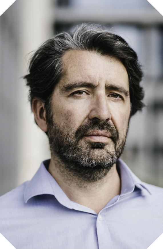 Portrait of a middle-aged man with dark hair and a beard, wearing a light blue collared shirt, standing outdoors with a blurred building background.
