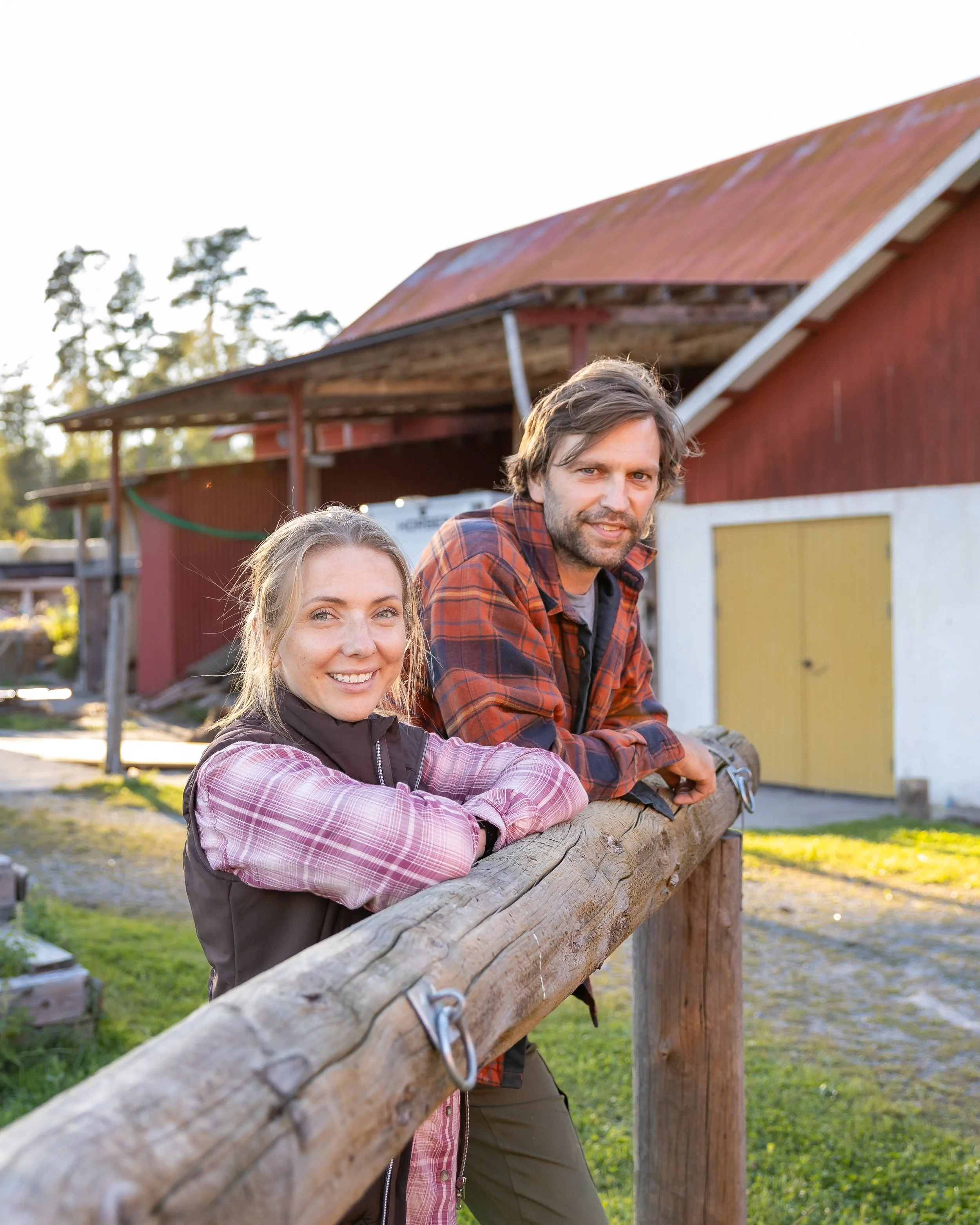 A smiling woman and man leaning on a wooden railing outdoors with a red barn in the background.