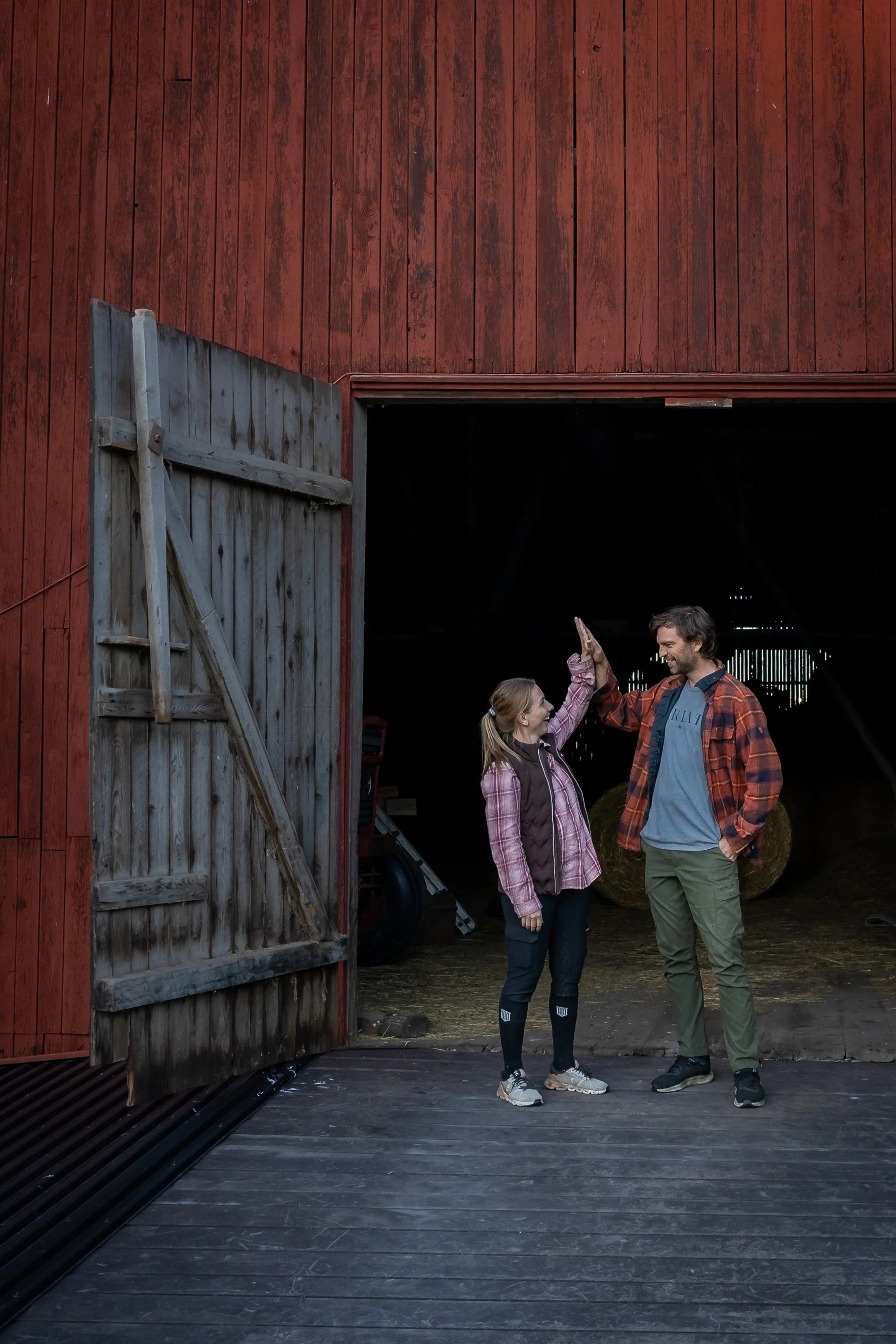 A man and woman giving each other a high five in front of a red barn, with hay bales inside.