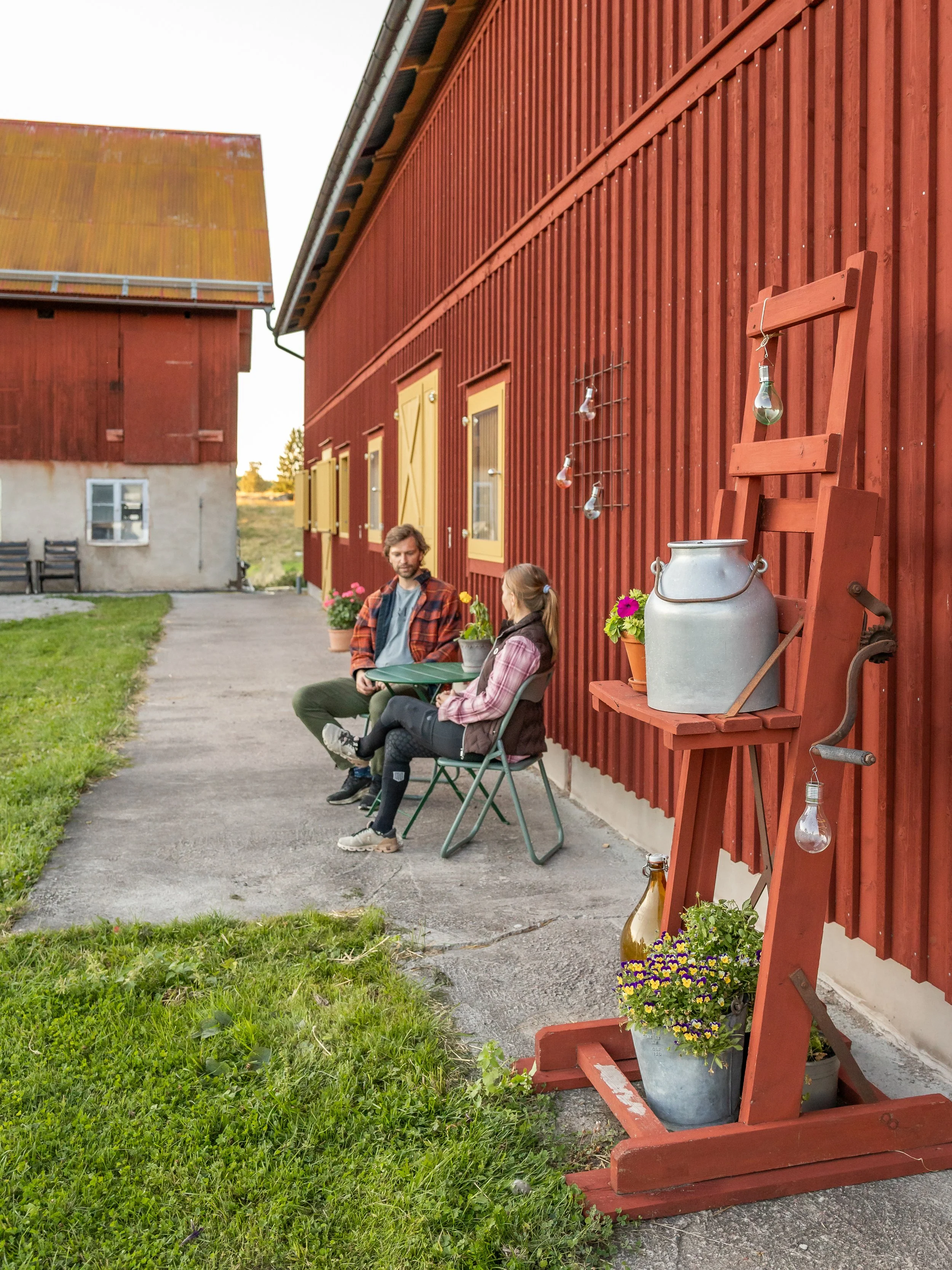 A man and woman sitting outside on folding chairs next to a red wooden building, having a conversation.