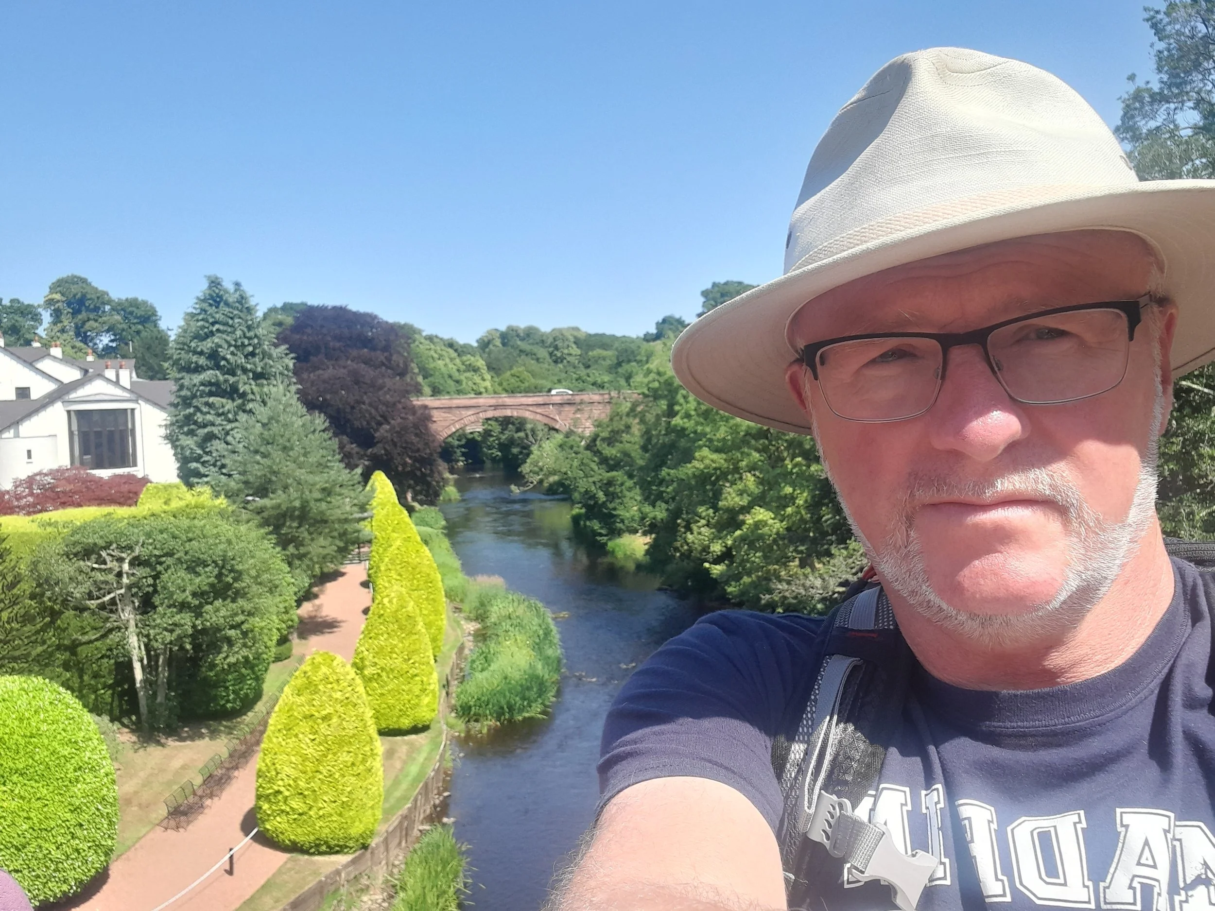A man wearing a wide-brim hat and glasses taking a selfie on a bridge with lush green trees and a river visible behind him.