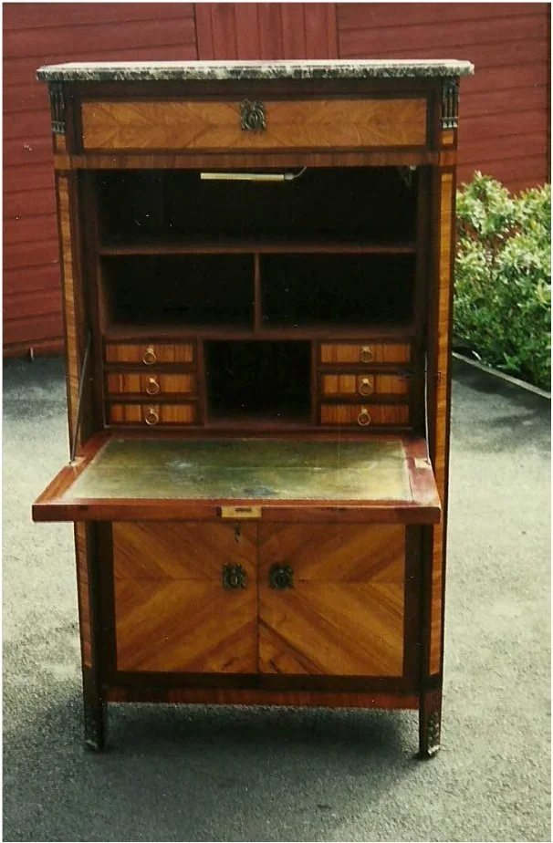 Antique wooden secretary desk with a marble top and small drawers, placed outdoors on asphalt with a red wooden fence and greenery in the background.