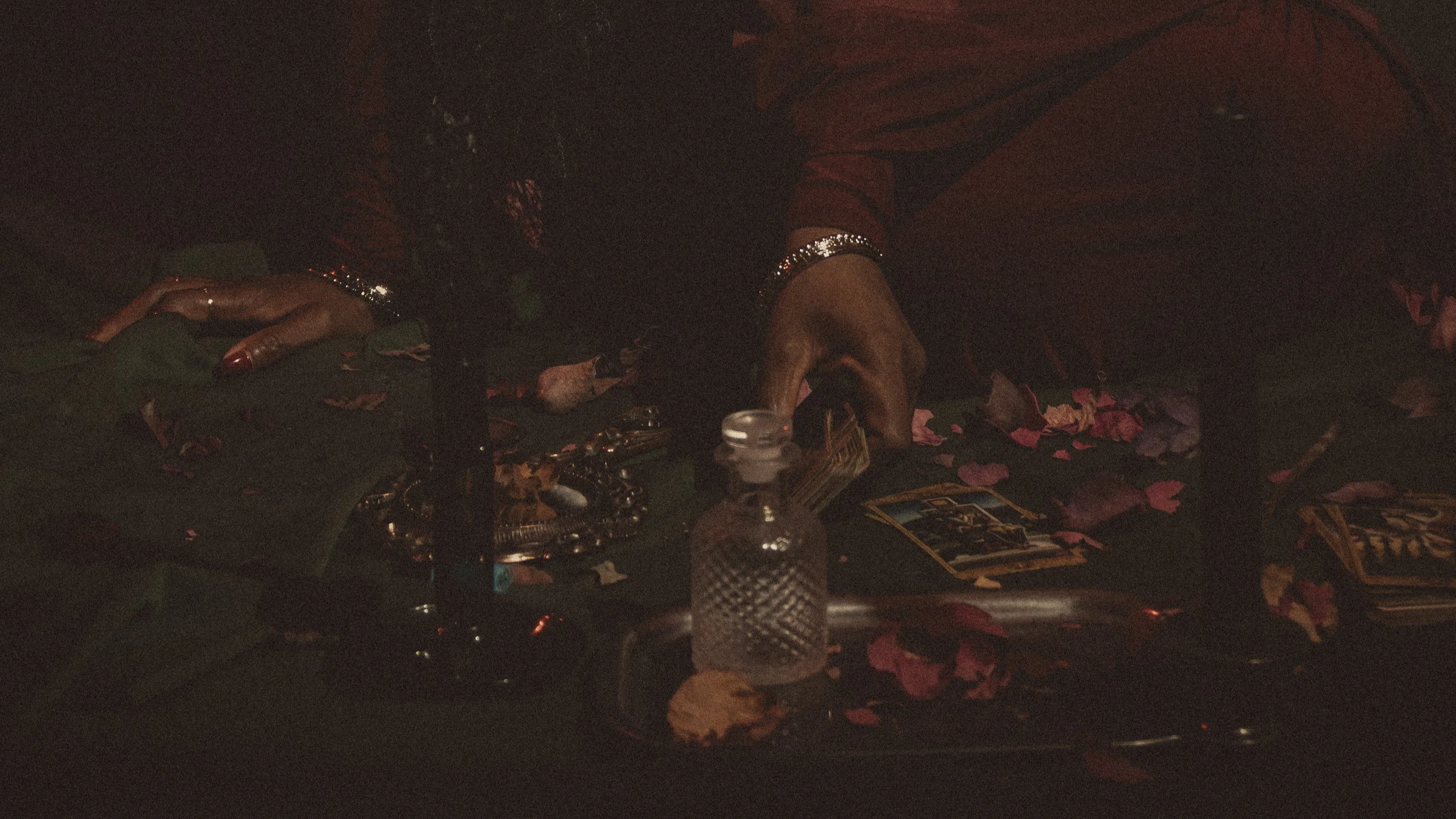 A woman dressed in deep red sits in a dimly lit ritual space surrounded by rose petals, tarot cards, and candlelight, evoking sacred feminine power and introspection