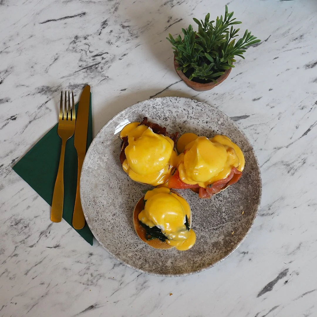 Plate of eggs Benedict with hollandaise sauce, served on a speckled ceramic plate, with a small potted plant nearby, on a white marble table, with green and gold utensils on a green napkin.