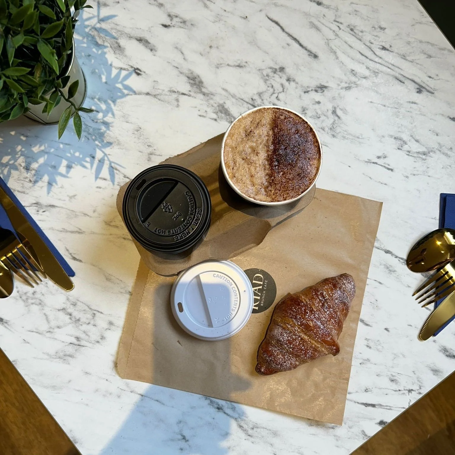 A breakfast spread on a white marble table, including a cup of cappuccino with frothy milk, a croissant dusted with powdered sugar, and two sealed coffee cups, one black and one white, placed on a brown paper napkin. There are gold utensils on either