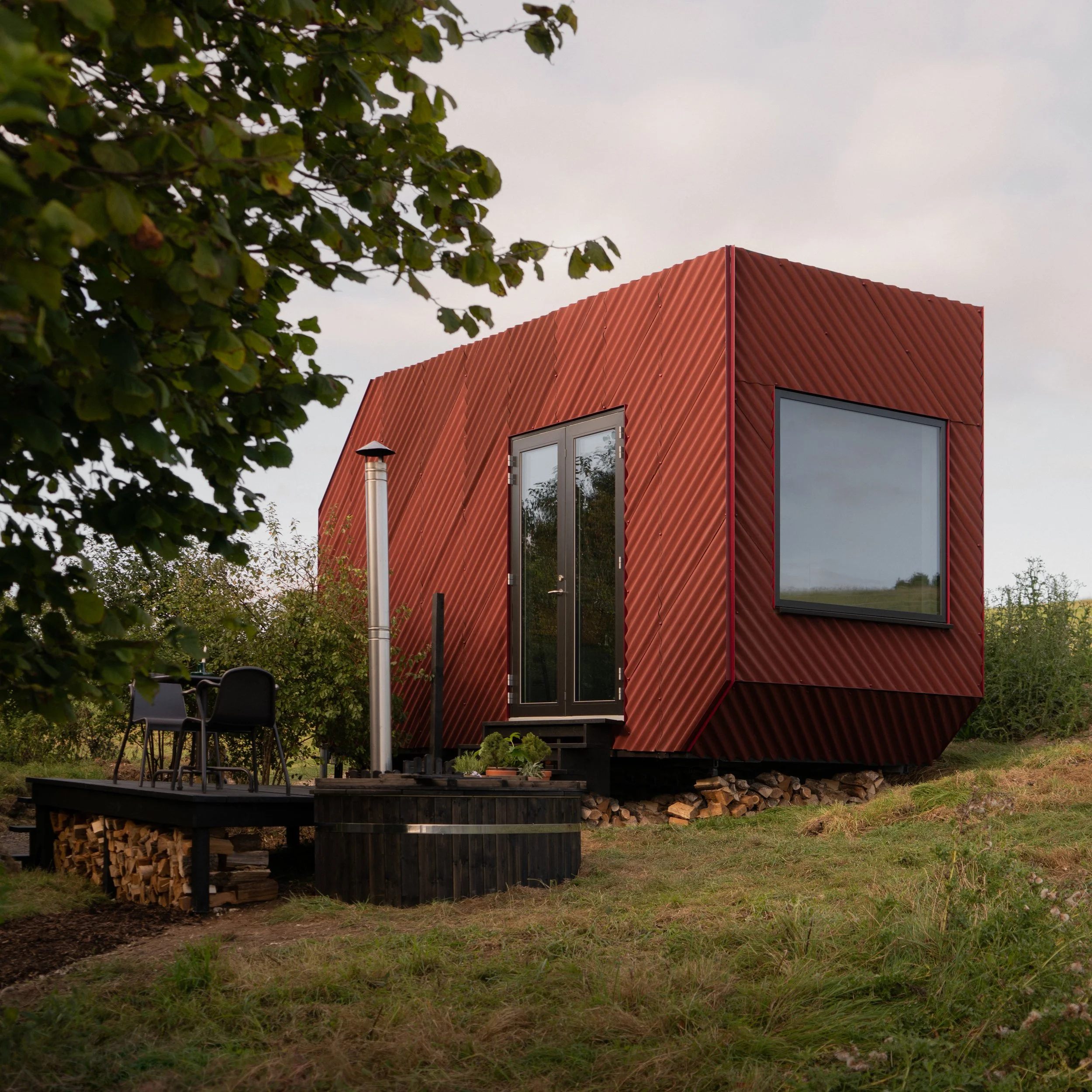 ReFrame Studio — A small, modern tiny house with a red, corrugated metal exterior, large windows, and a set of double doors, situated on a grassy area with a small out