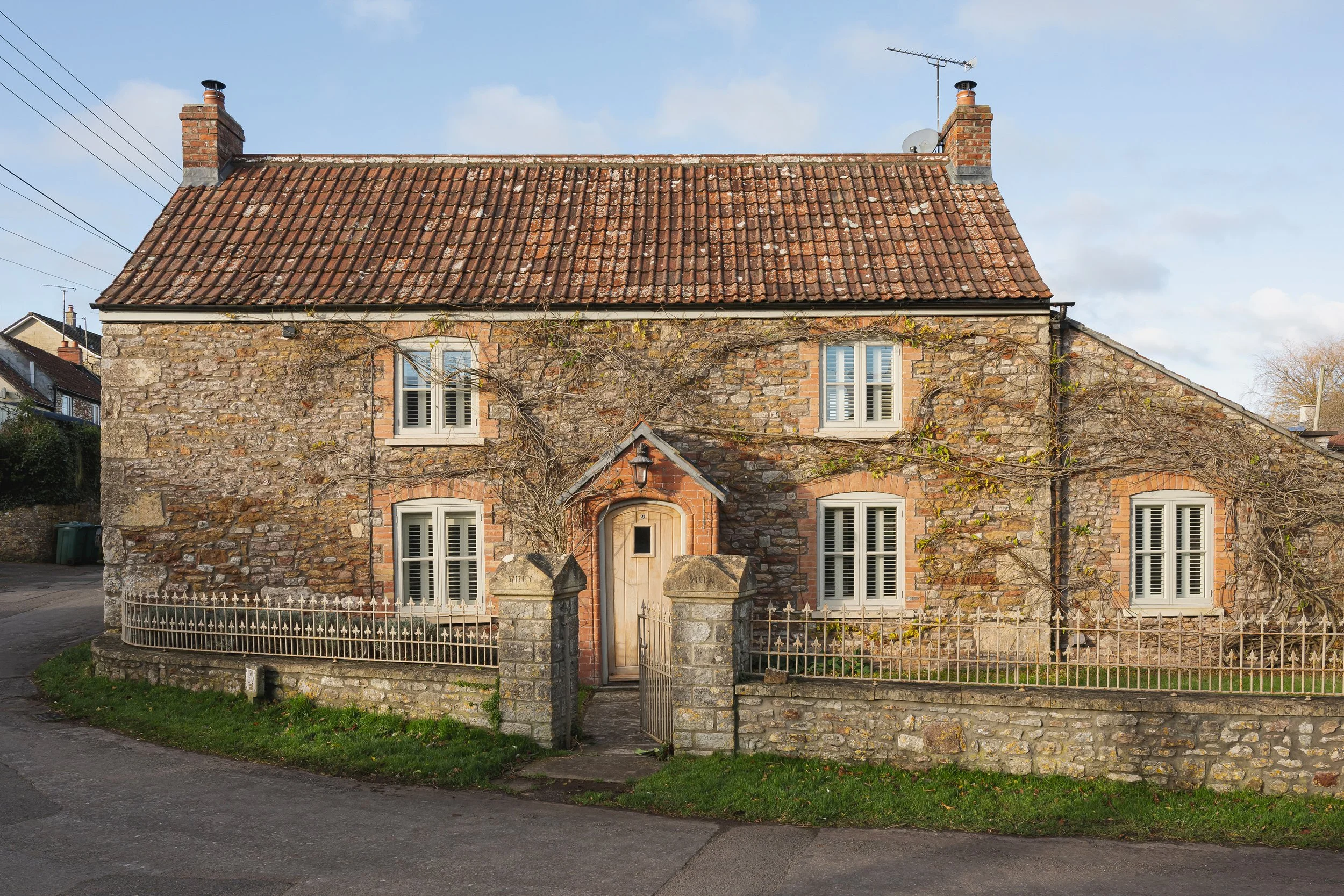 ReFrame Studio — A two-story stone house with a tiled steep roof, two chimneys, and six window shutters, surrounded by a stone and metal fence with a small gate.