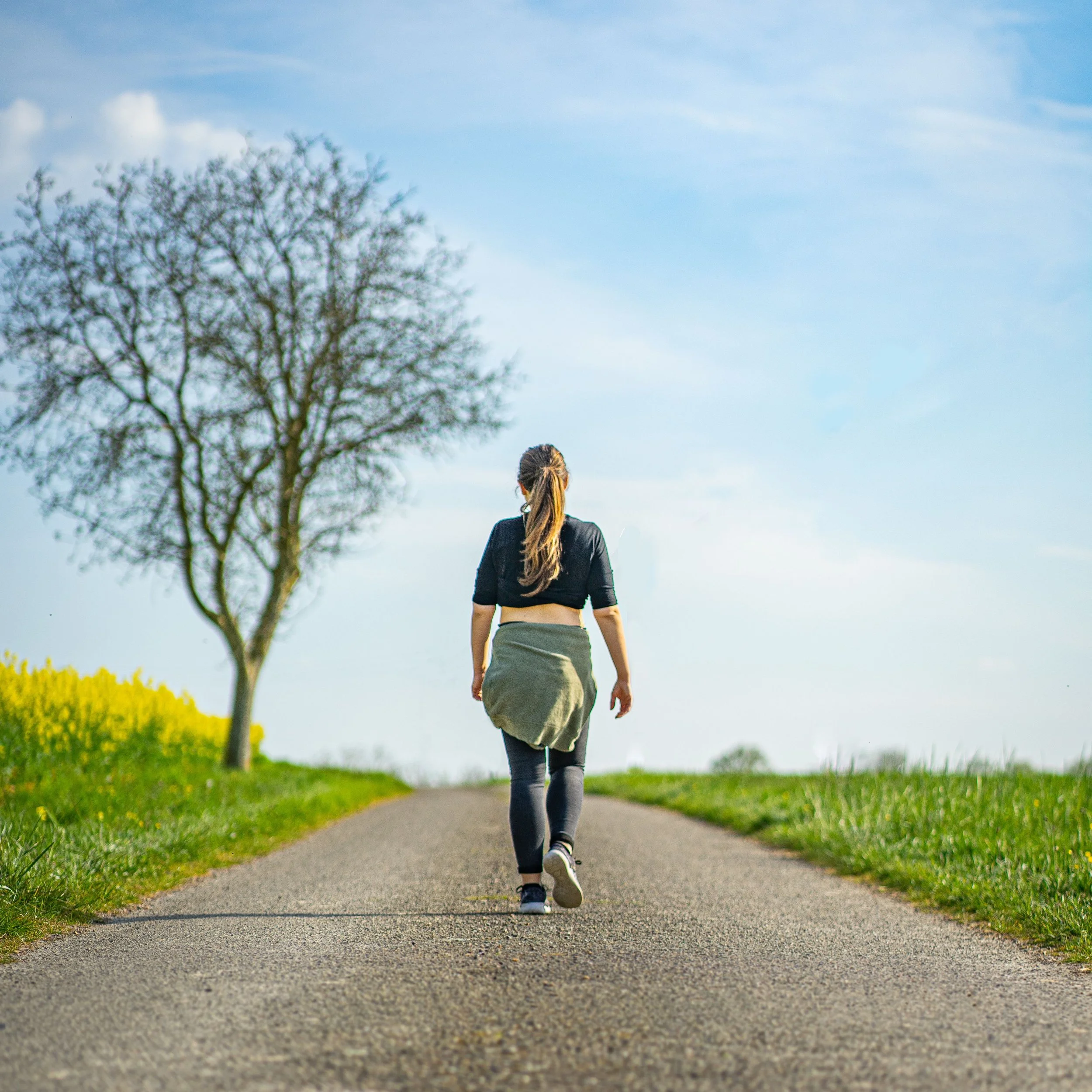 A woman walking down a road with bright green grass on both the left and right side with spring yellow flowers growing in the field on the left.