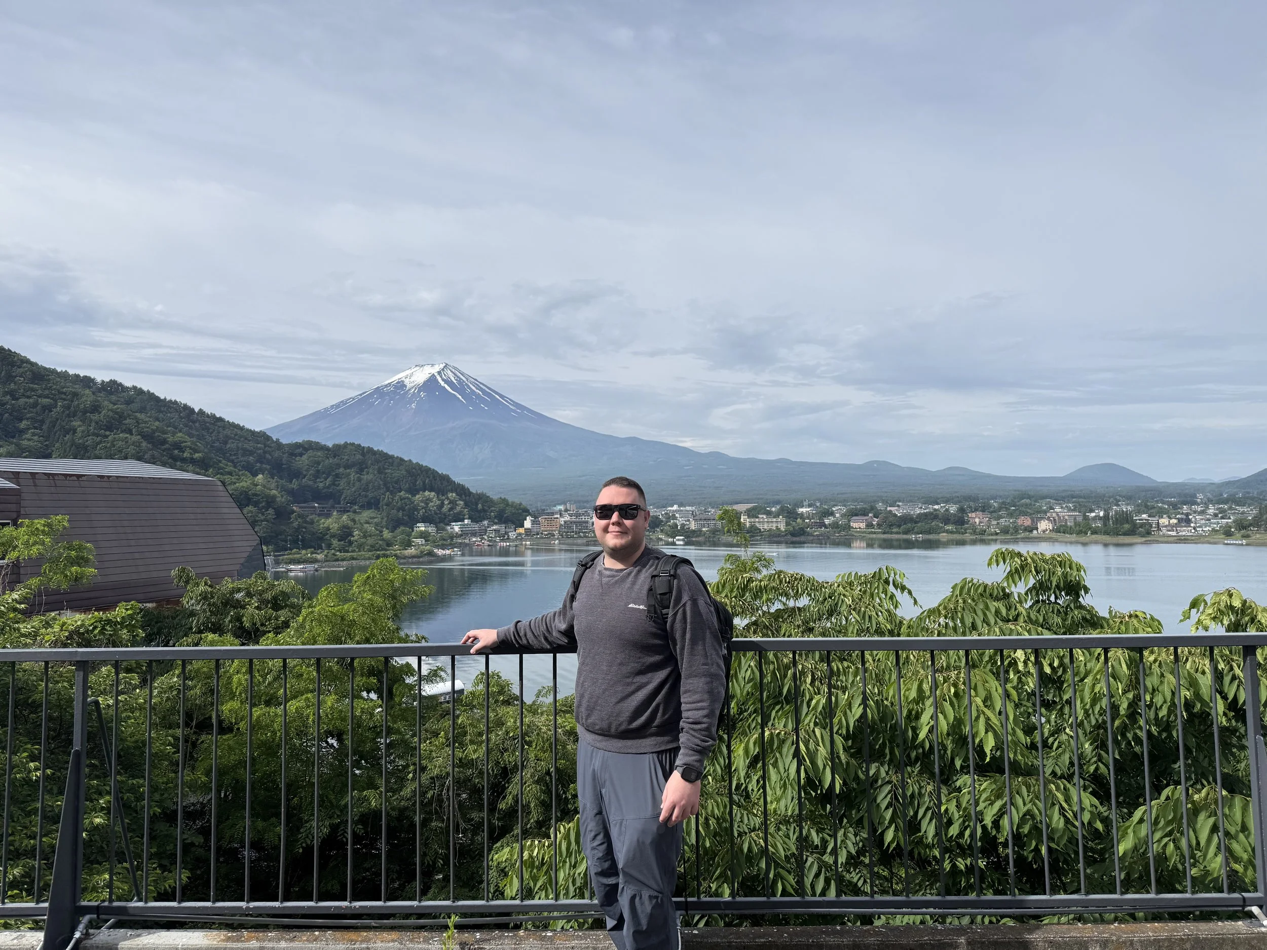 A man with sunglasses and a backpack standing by a railing, with Mount Fuji and a river in the background, under a partly cloudy sky.