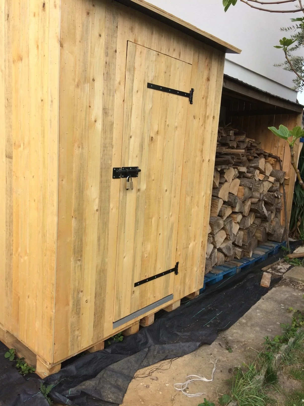 Une petite cabane en bois avec une serrure à cadenas, située dans un jardin, avec un tas de bois empilé à côté.