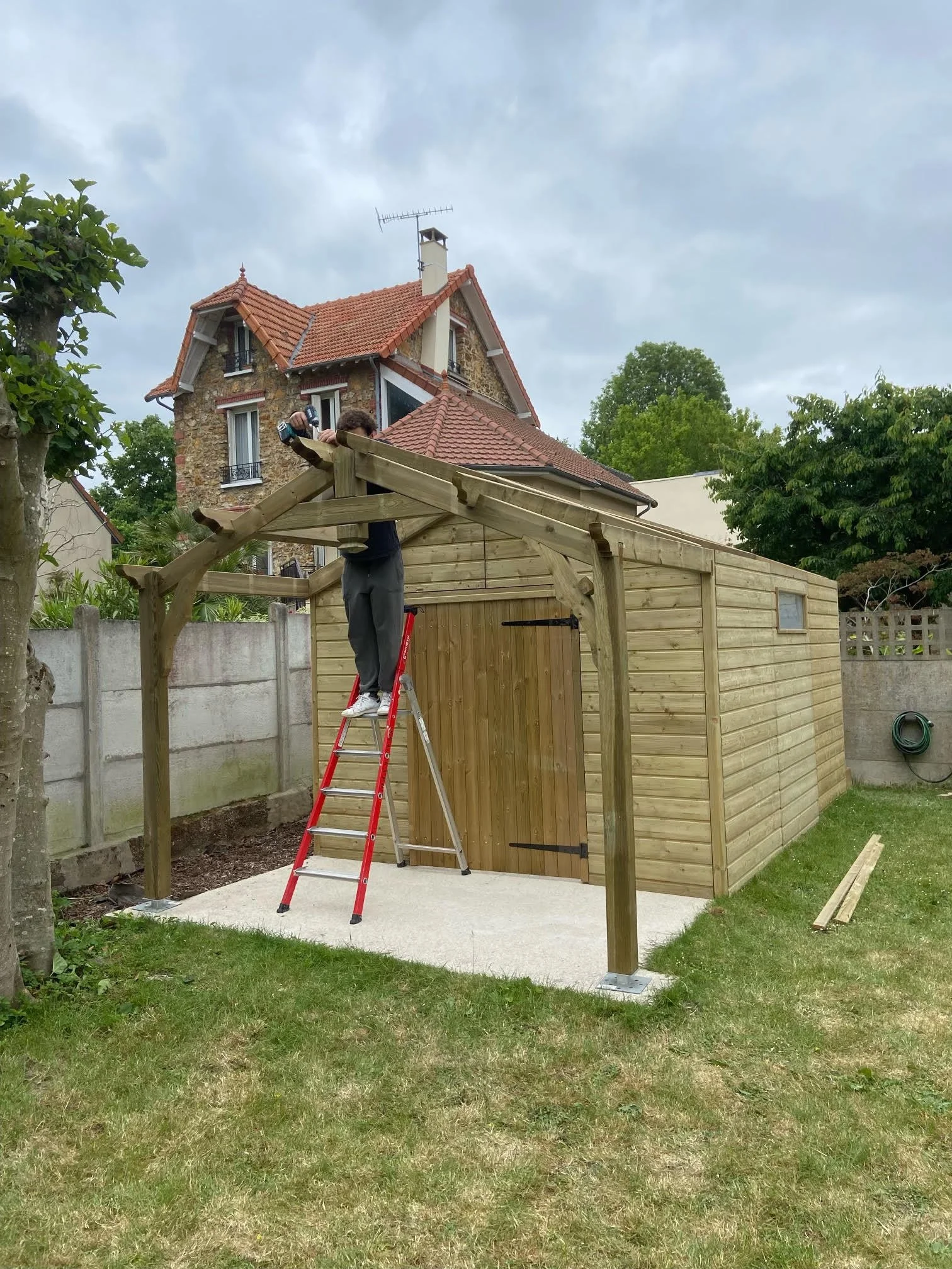Une personne construisant une petite structure en bois dans un jardin, avec une échelle rouge et un plancher en béton. Un grand maison en pierre avec un toit en tuiles rouges est visible en arrière-plan, entourée d'arbres verts et un ciel nuageux.