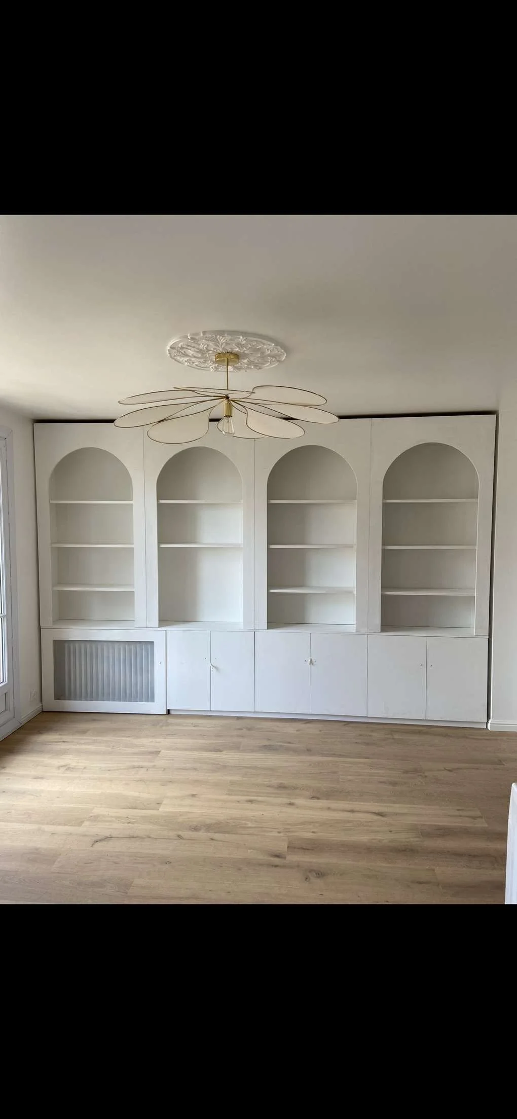 Une étagère blanche encastrée avec des portes en bas dans une pièce avec un plafond blanc, un lustre en forme de fleur, et un sol en bois clair.