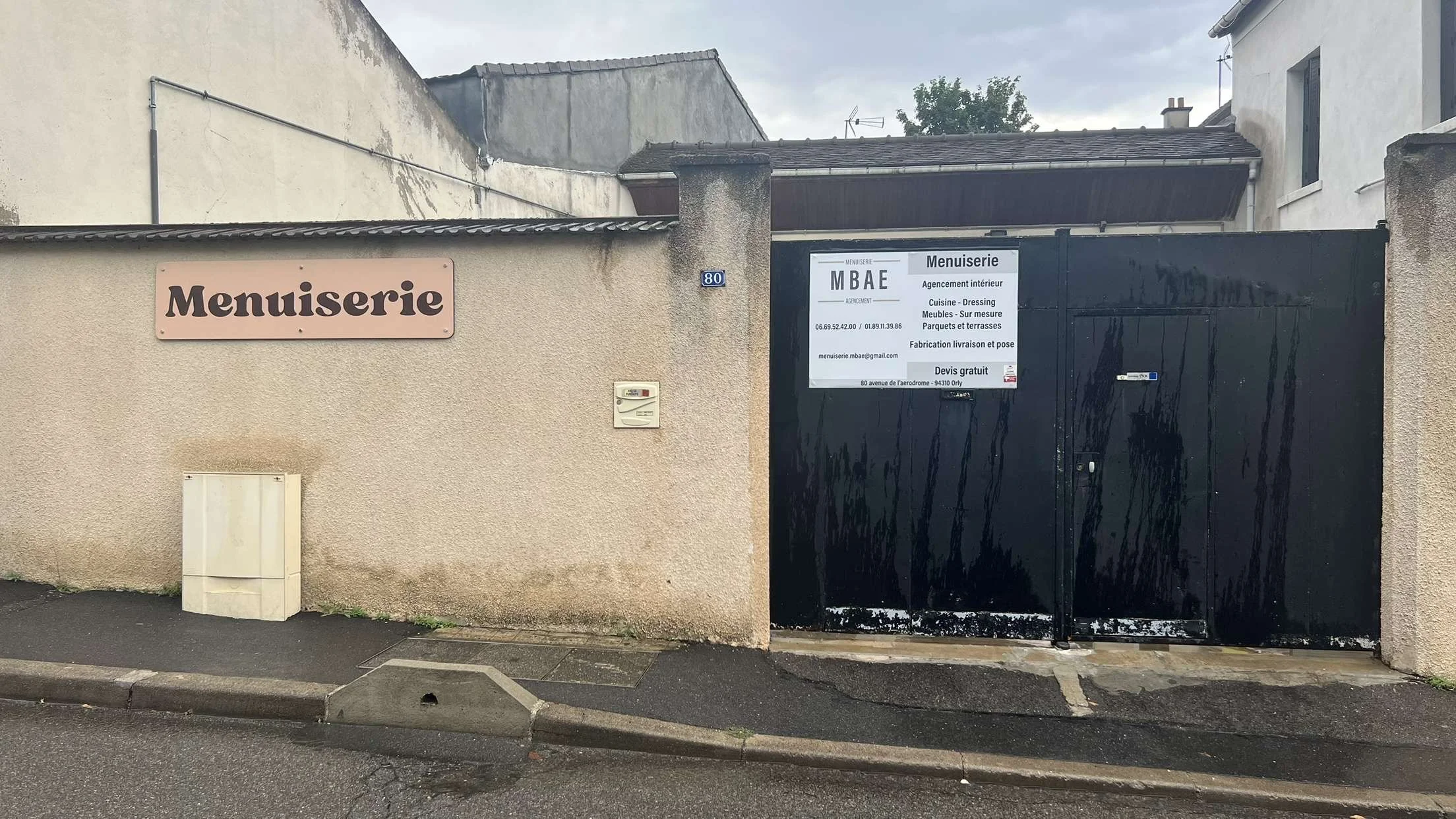 Une porte noire avec un panneau publicitaire et un panneau indiquant l'adresse, situé derrière un mur beige. La rue en avant est pavée, avec une ligne de stationnement au bord.