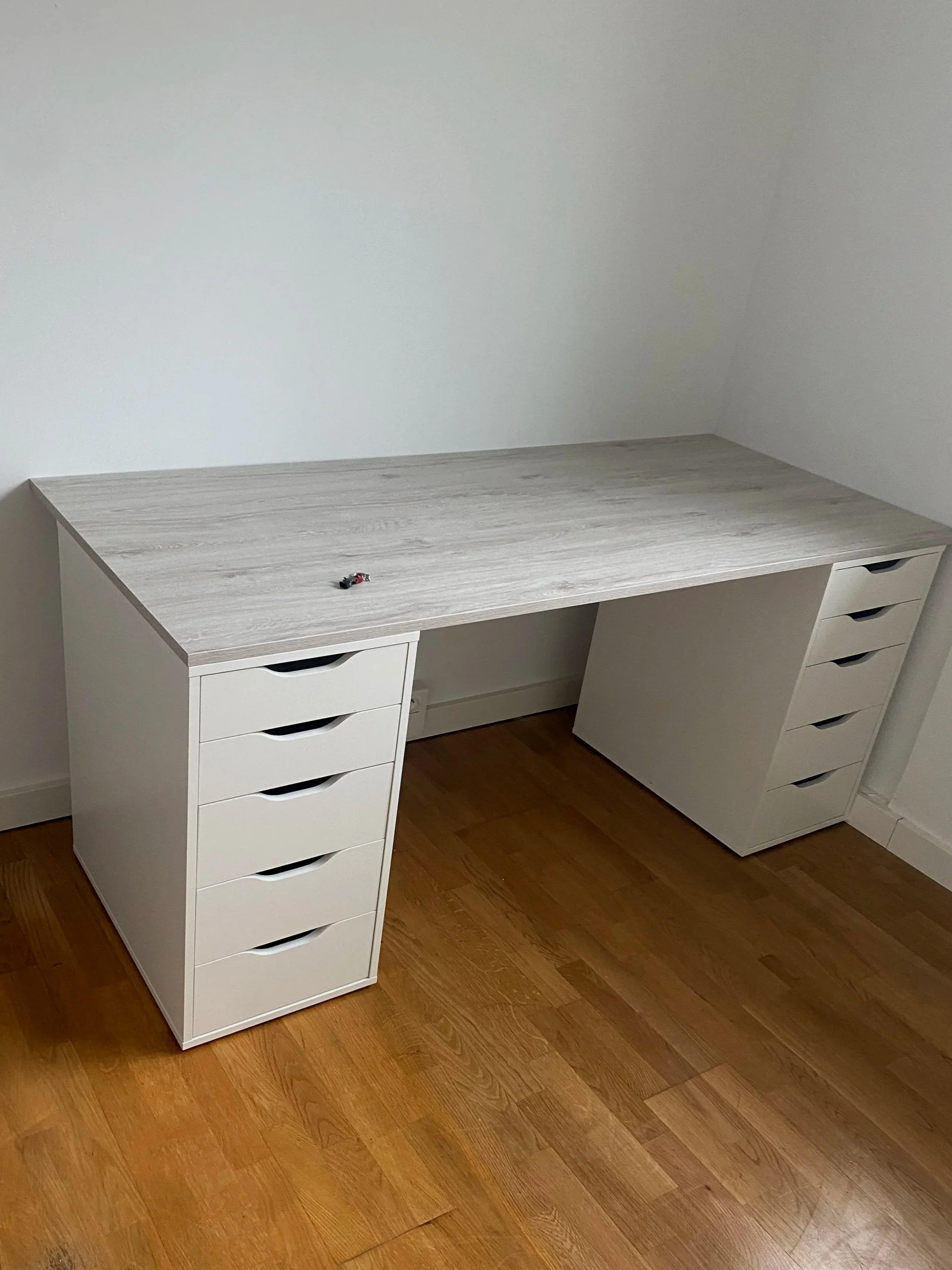 Un bureau en bois clair et blanc avec plusieurs tiroirs blancs, placé dans une pièce avec un sol en parquet de bois foncé et un mur blanc vide en arrière-plan.