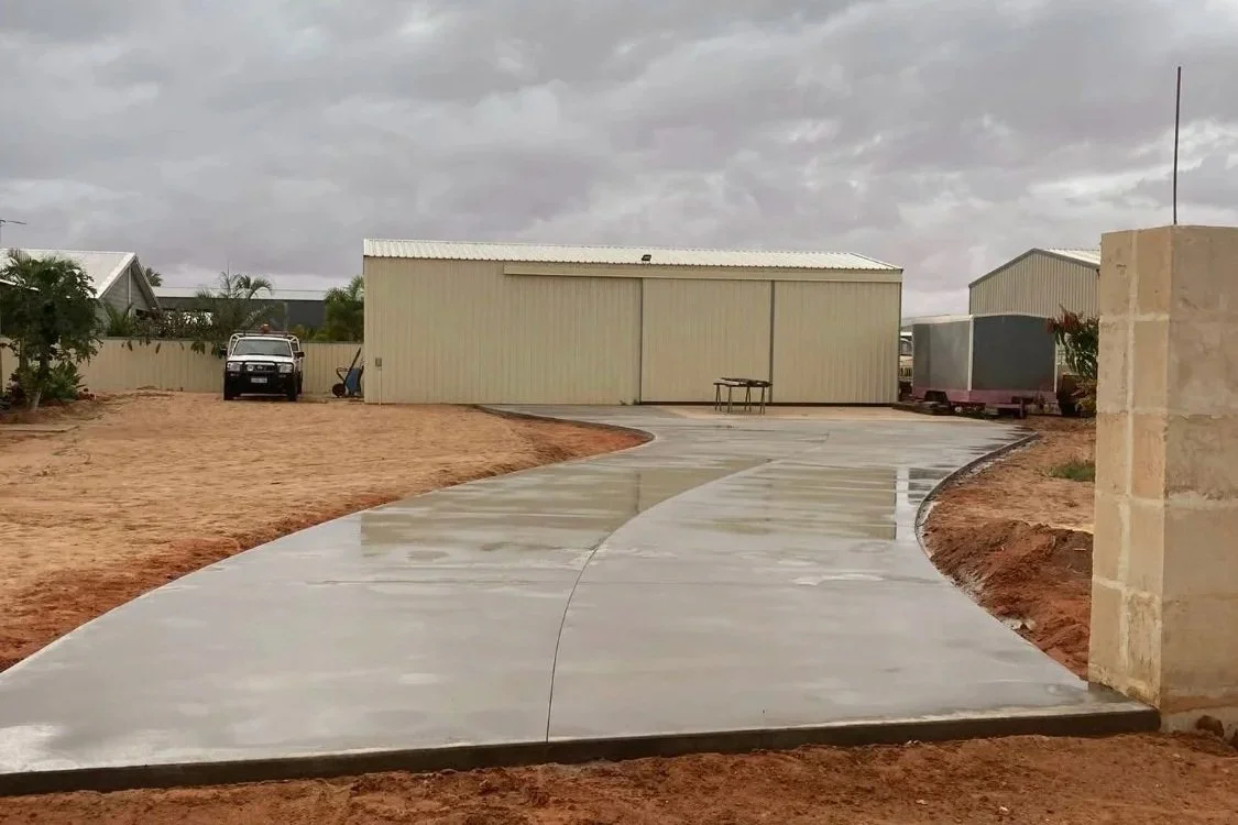 Concrete sidewalk with a curved pathway leading to a storage shed in a yard, with a car parked to the left, cloudy sky, and some plants and structures in the background.