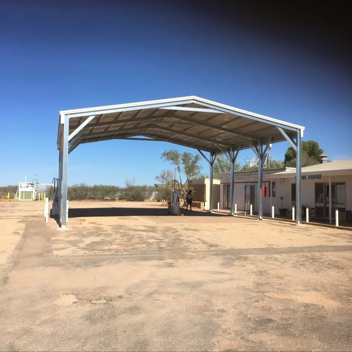 A metal carport structure with twin fuel pumps underneath. The area appears to be a small, remote gas station or rest stop with a building nearby and a dirt ground. Clear blue sky in the background.