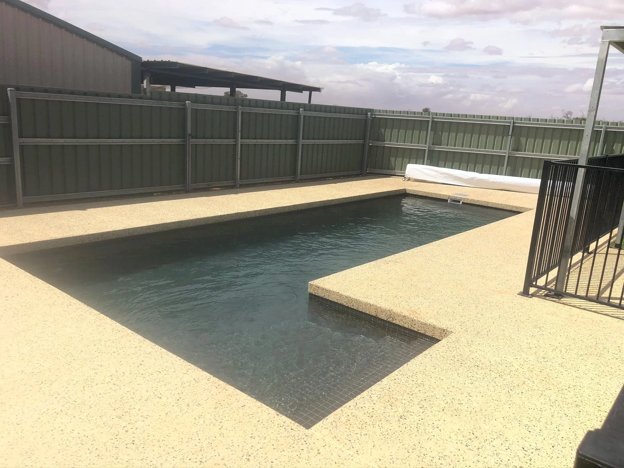 Small rectangular backyard pool with a hot tub attached, surrounded by beige textured concrete, with a green metal fence and cloudy sky in the background.