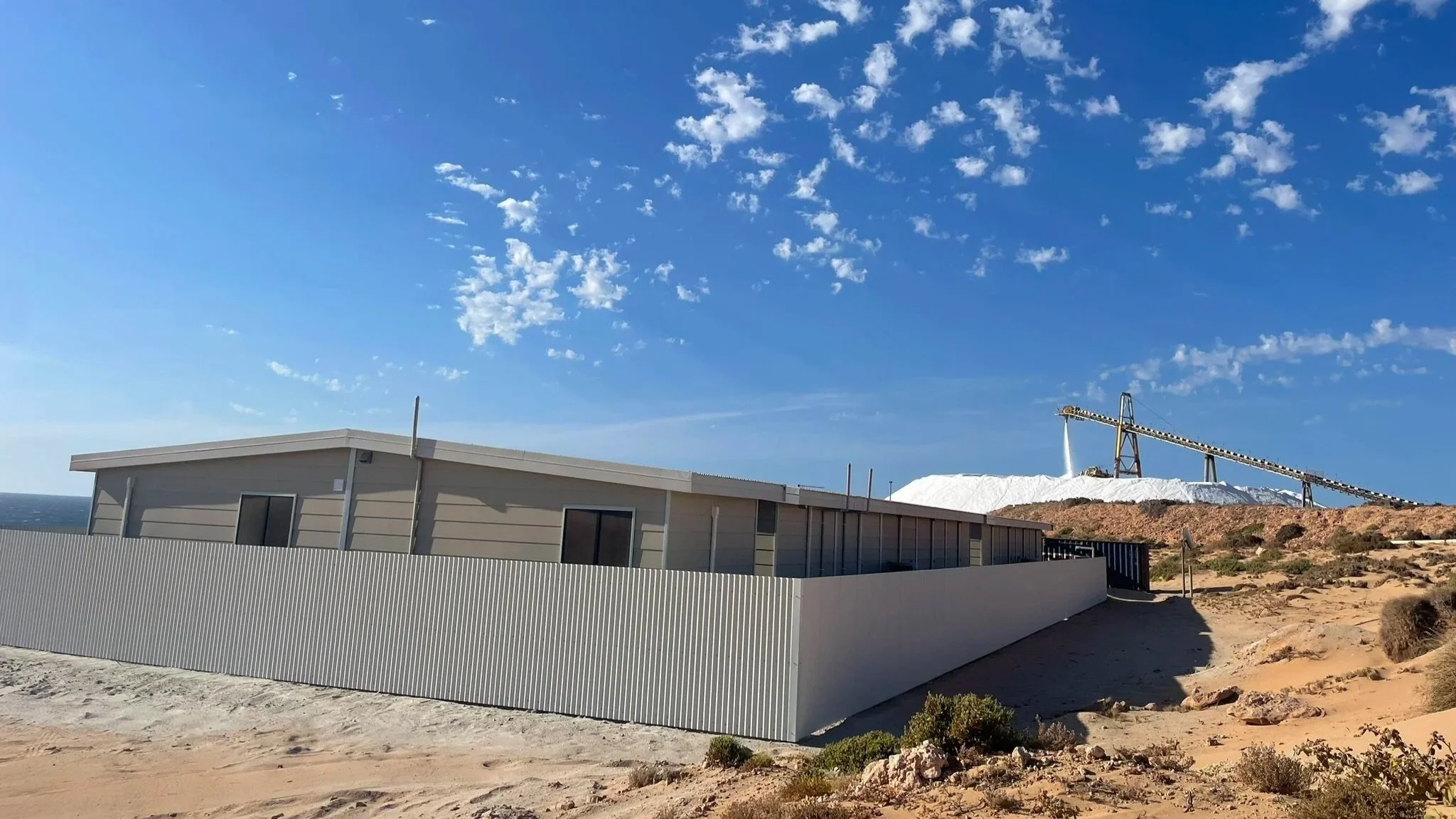 A modern, single-story building with corrugated metal walls set on sandy terrain, with a salt harvesting pond and conveyor system in the background under a blue sky with scattered clouds.