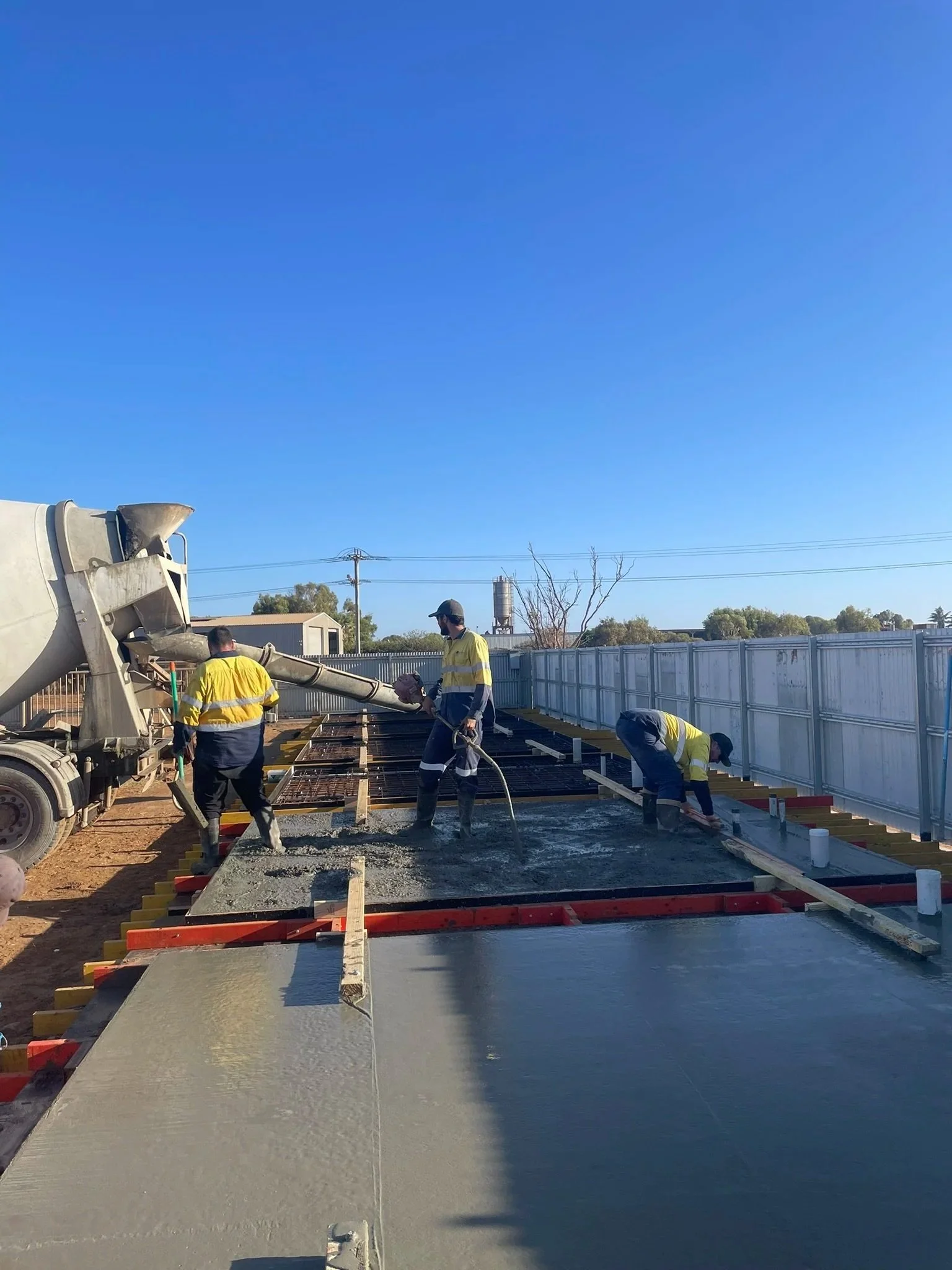 Workers in safety vests and helmets pouring and leveling concrete for a building foundation outside under a clear blue sky.