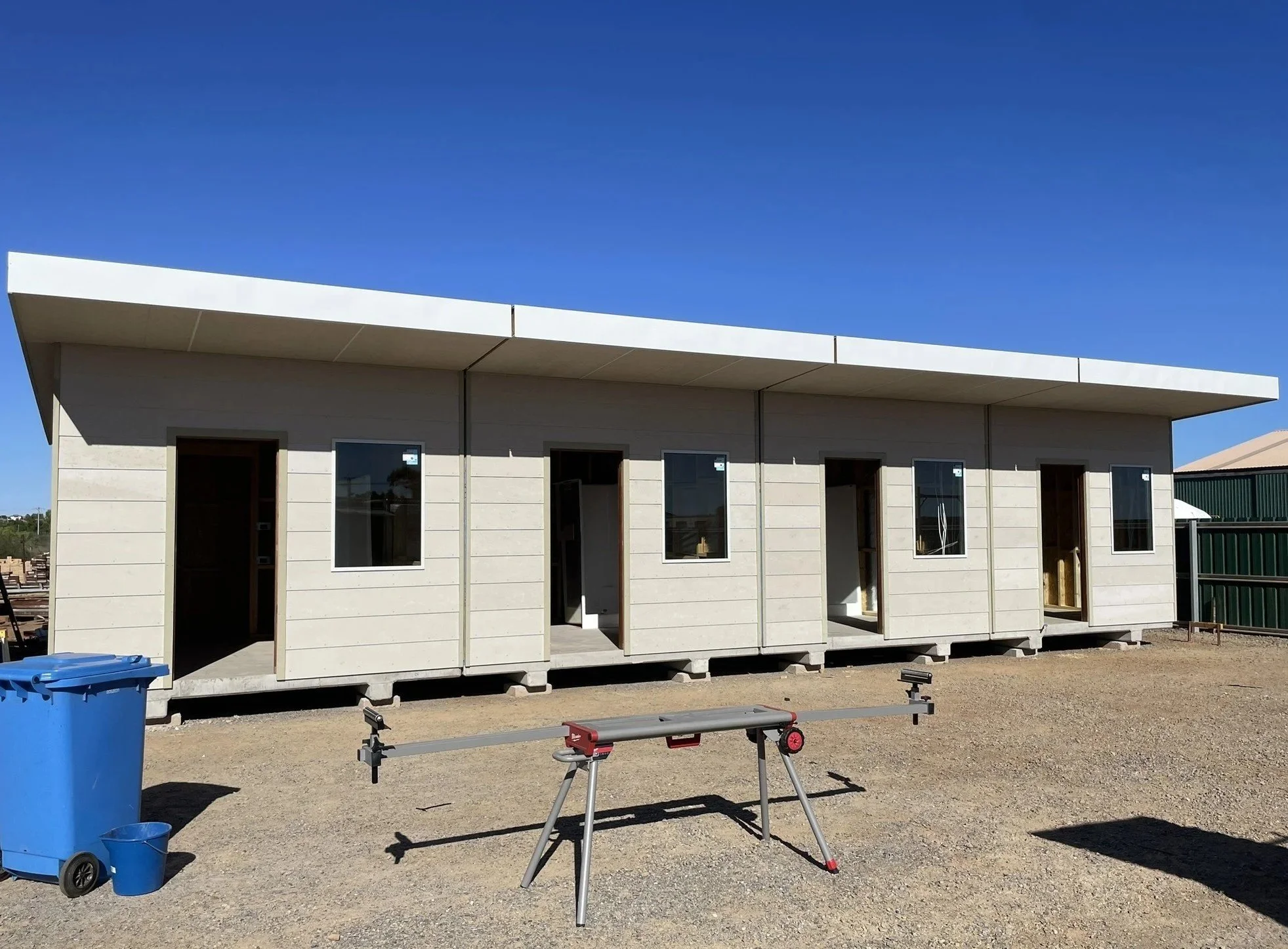 Under construction tiny house with three open doorways and four windows, set on a gravel lot with a blue trash bin and a workbench in the foreground, against a clear blue sky.