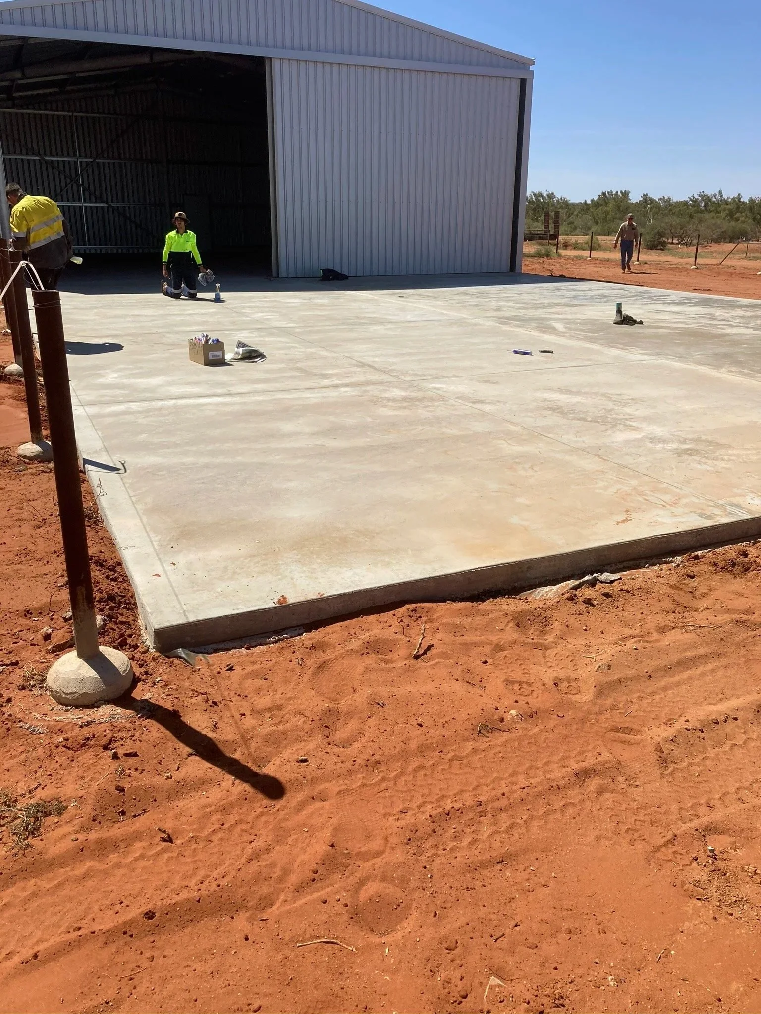 Concrete slab in front of a metal building under construction with workers and tools present, in a dry, red dirt landscape.