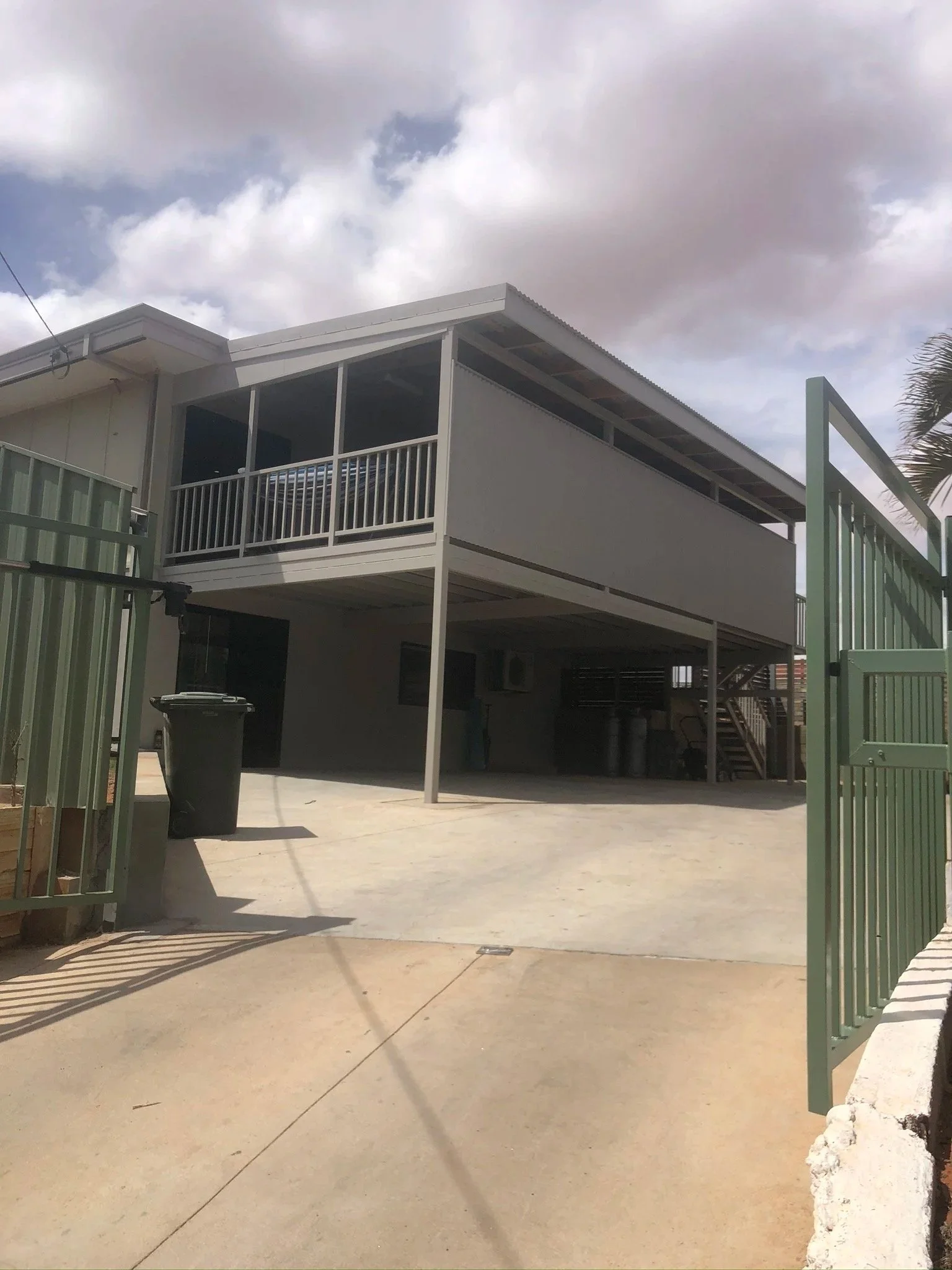 A two-story house with a balcony and staircase, viewed from the driveway with a trash bin and a green gate in the front.