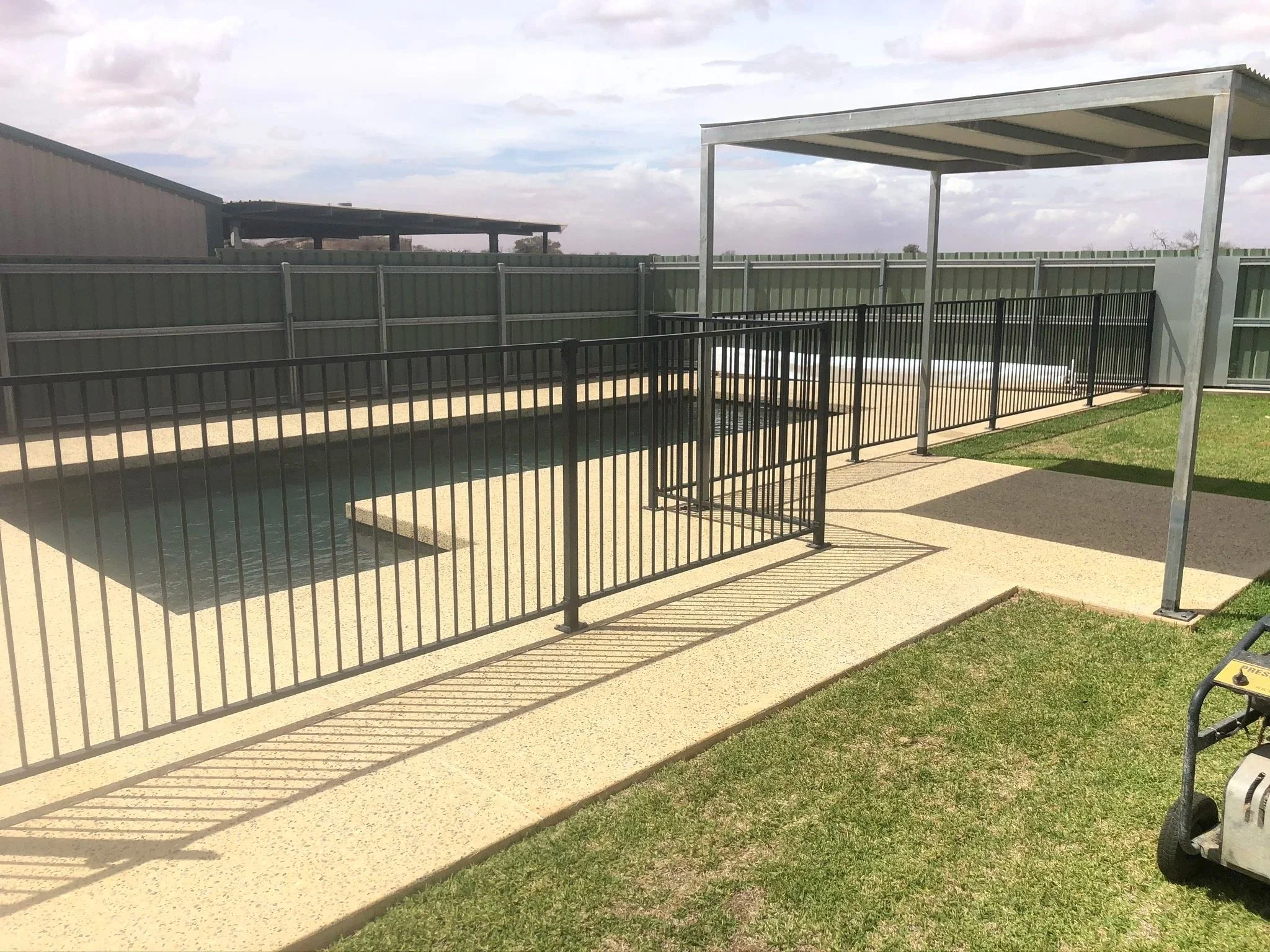 A fenced-in outdoor pool area with a small pool, black metal fences, a shaded structure, and a grassy area on a partly cloudy day.