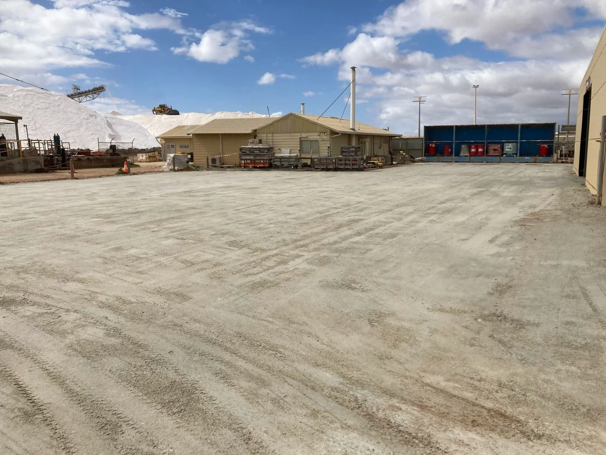 An empty industrial yard with a large gravel surface, some buildings, equipment, and a large pile of salt or snow in the background under partly cloudy skies.