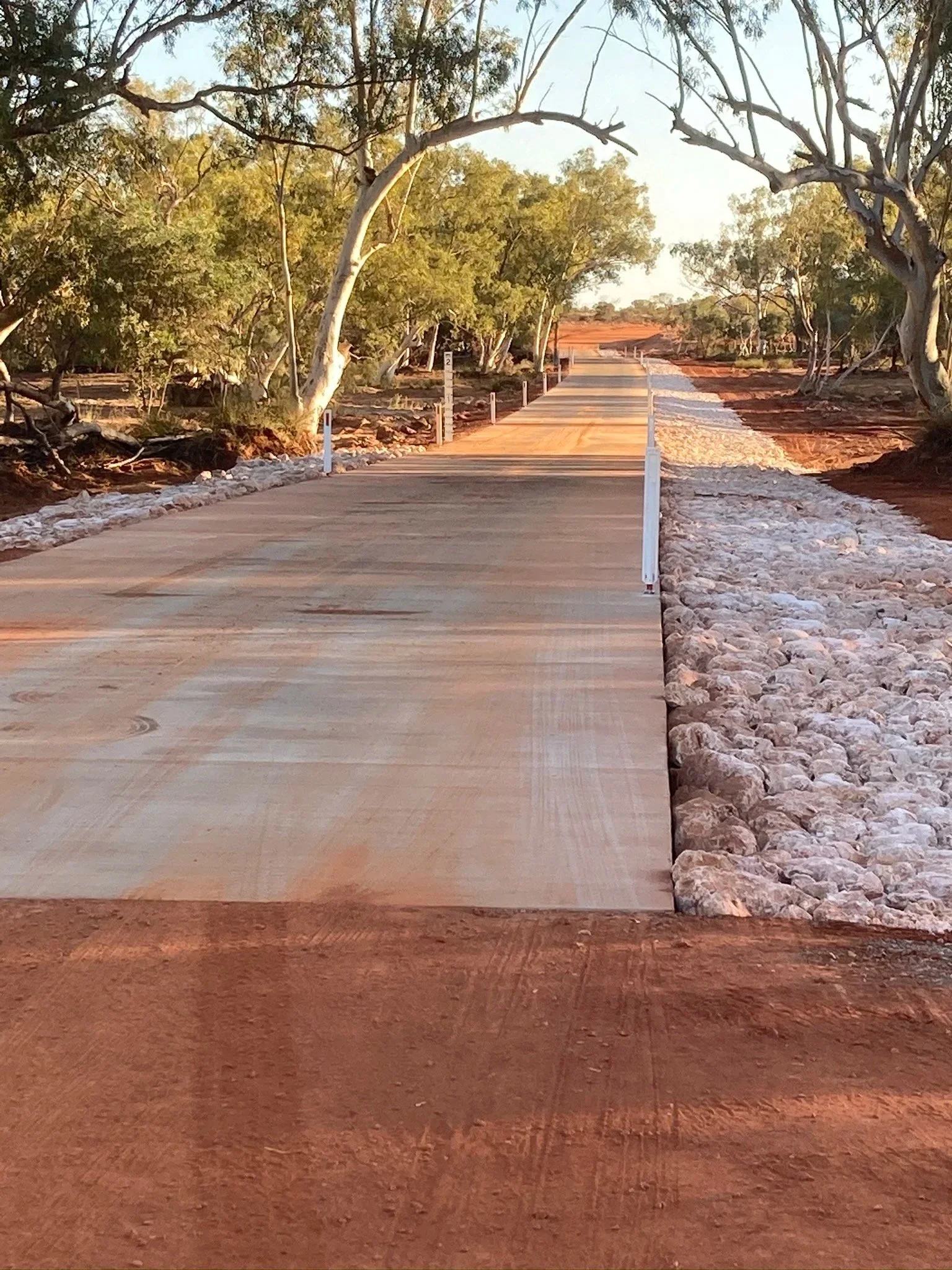 A newly constructed pathway with a concrete surface runs through a dry, wooded landscape, lined with small white posts on either side, and surrounded by red earth and white rocks.